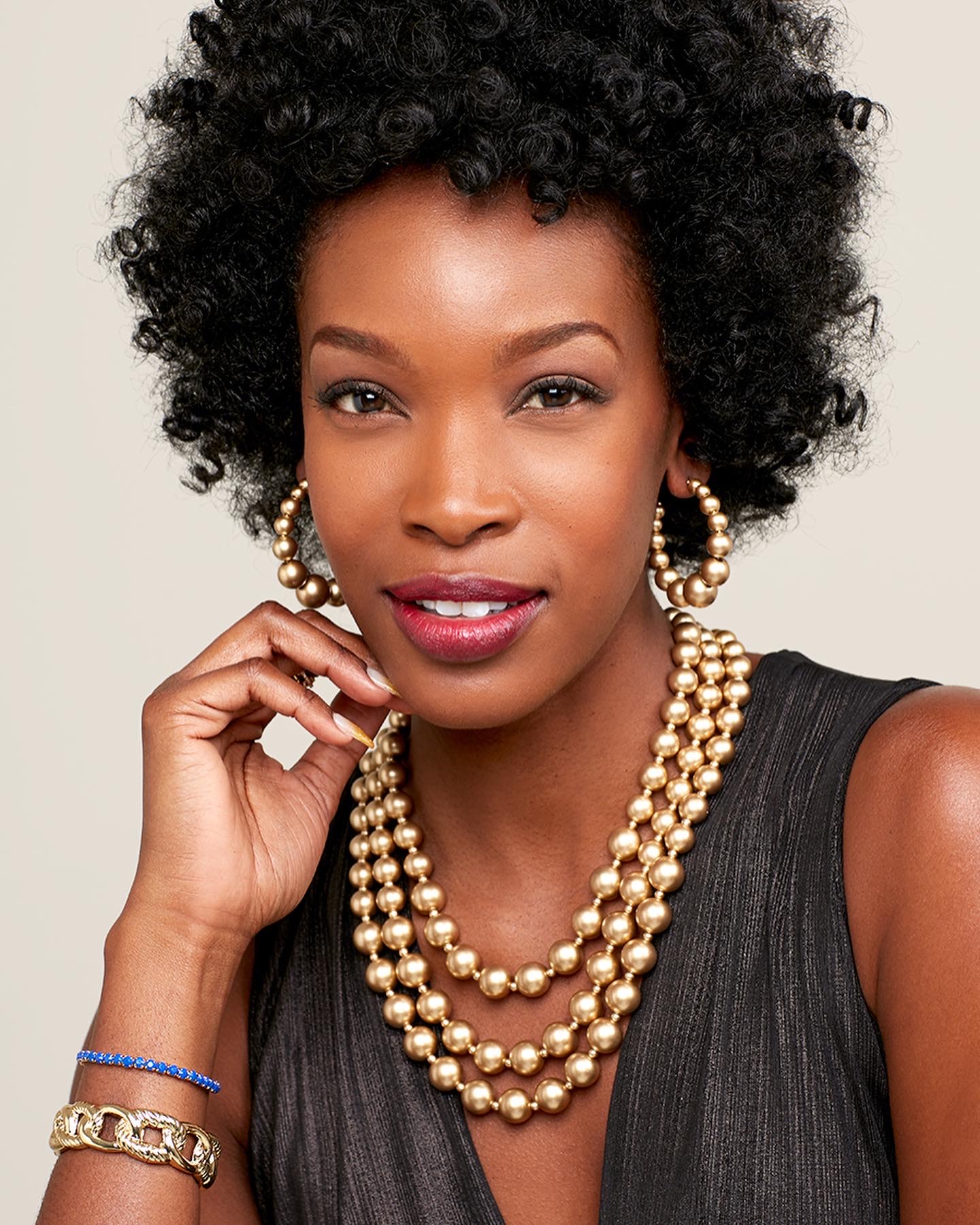 Portrait of a woman with curly hair wearing pearl necklaces, earrings, and a sleeveless dark top, smiling gently.