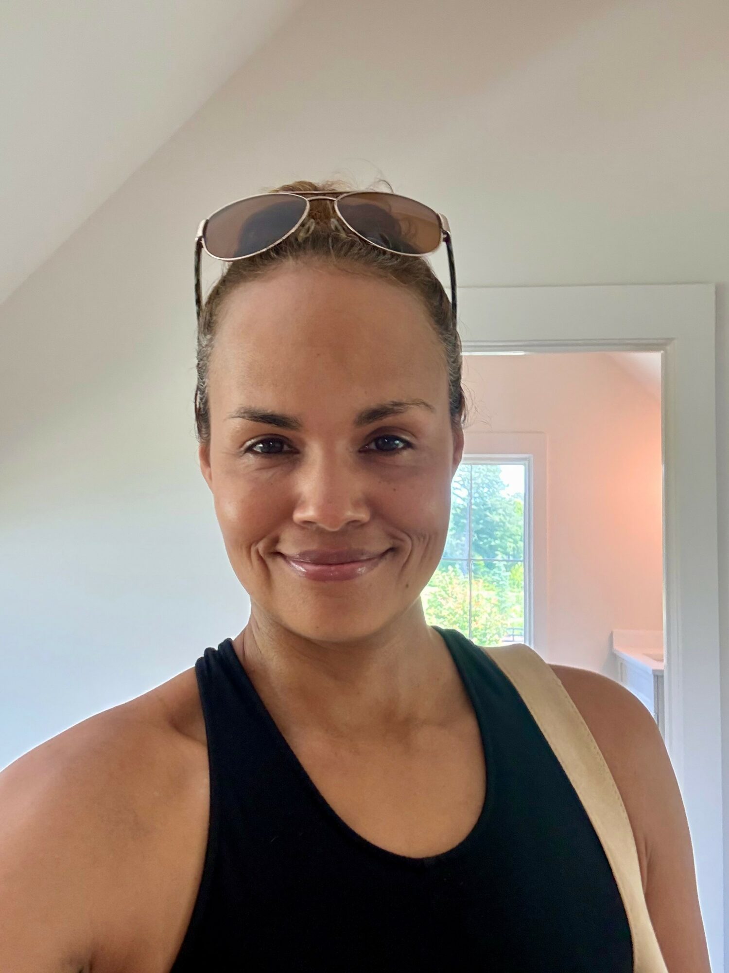 Woman with sunglasses on her head, smiling, wearing a black tank top, indoors near a window.