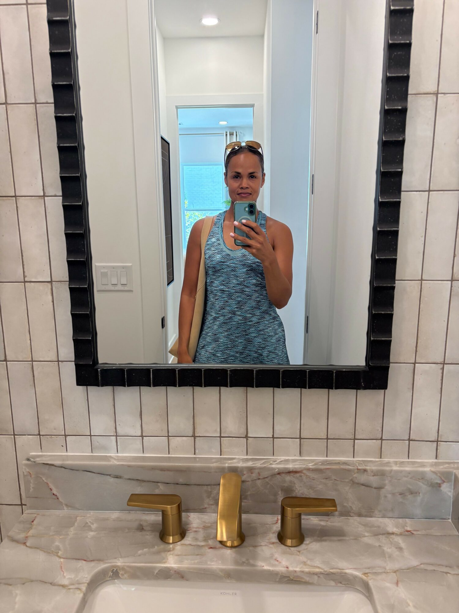 Person taking a mirror selfie in a bathroom, wearing a blue patterned dress, with a window in the background.