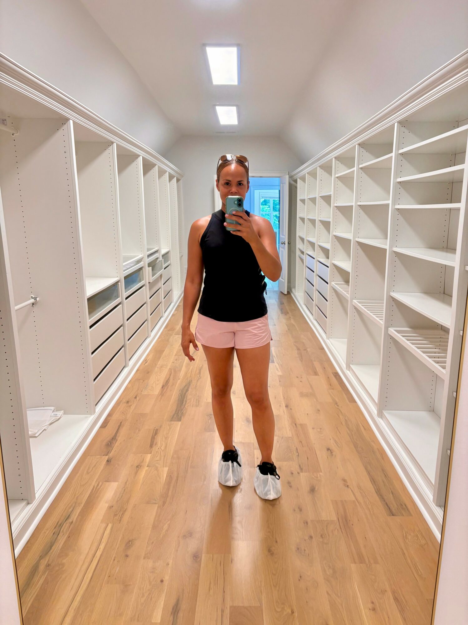 Person taking a mirror selfie in an empty store aisle with white shelves on both sides, wearing a black top and pink shorts.