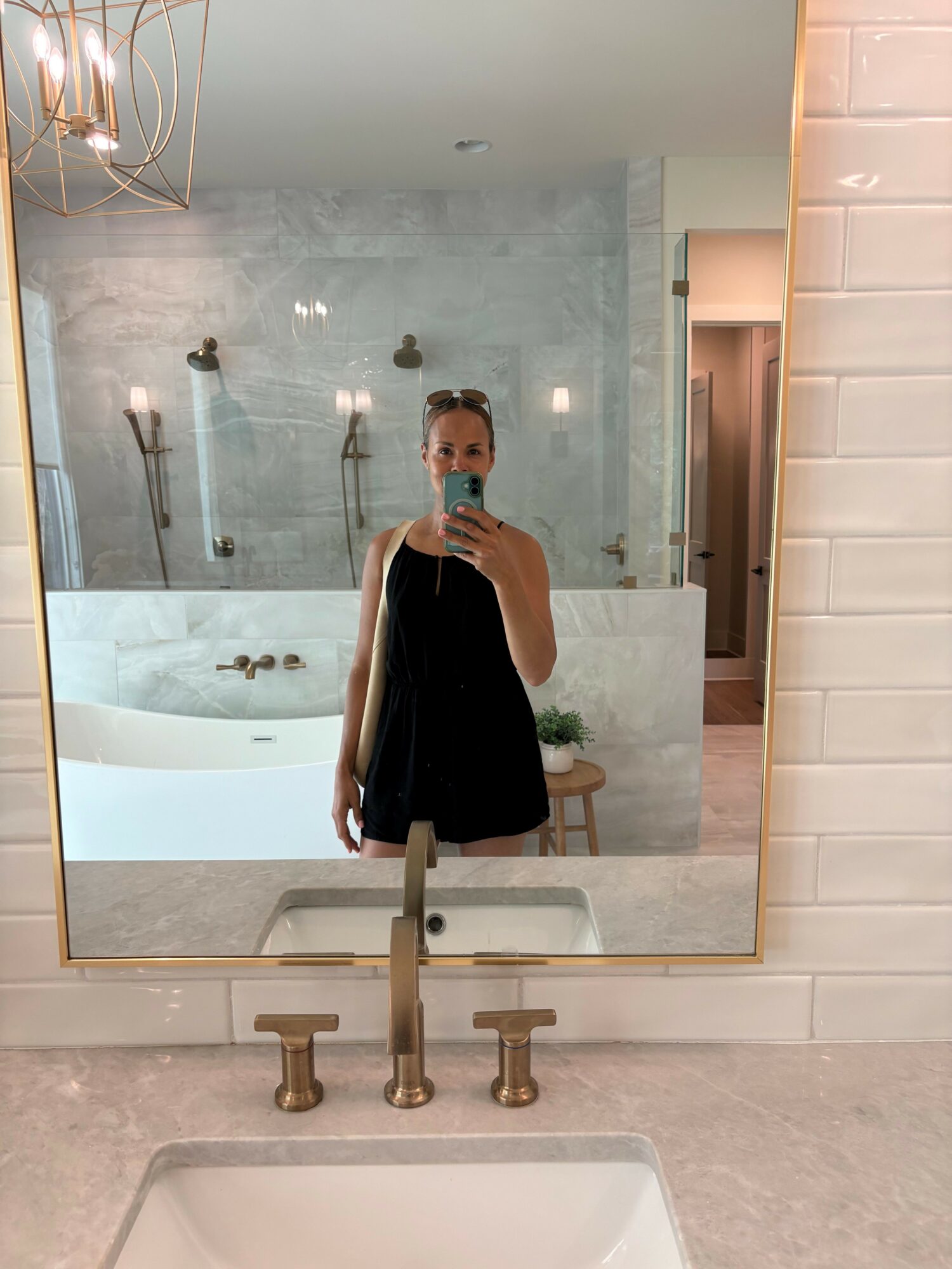 Person taking a mirror selfie in a bathroom with a shower, bathtub, and beige tiles, wearing a black dress.