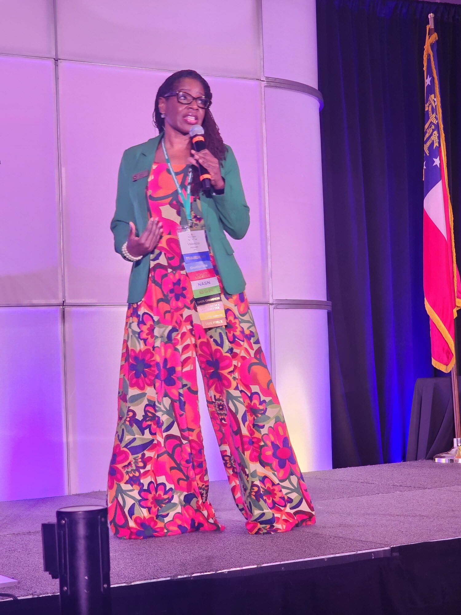 Woman standing on stage, speaking into microphone, wearing colorful floral pants and green blazer, with a flag in the background.