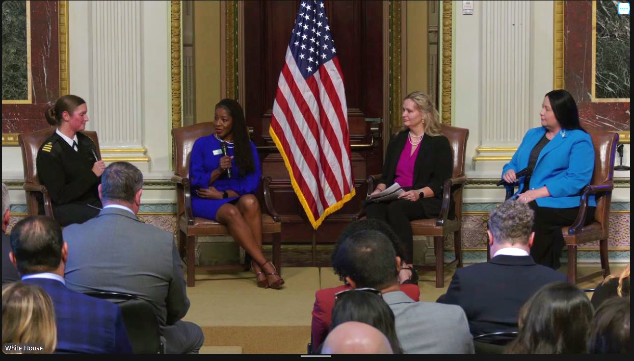Four women seated on stage in front of an American flag, speaking to an audience in a formal setting.