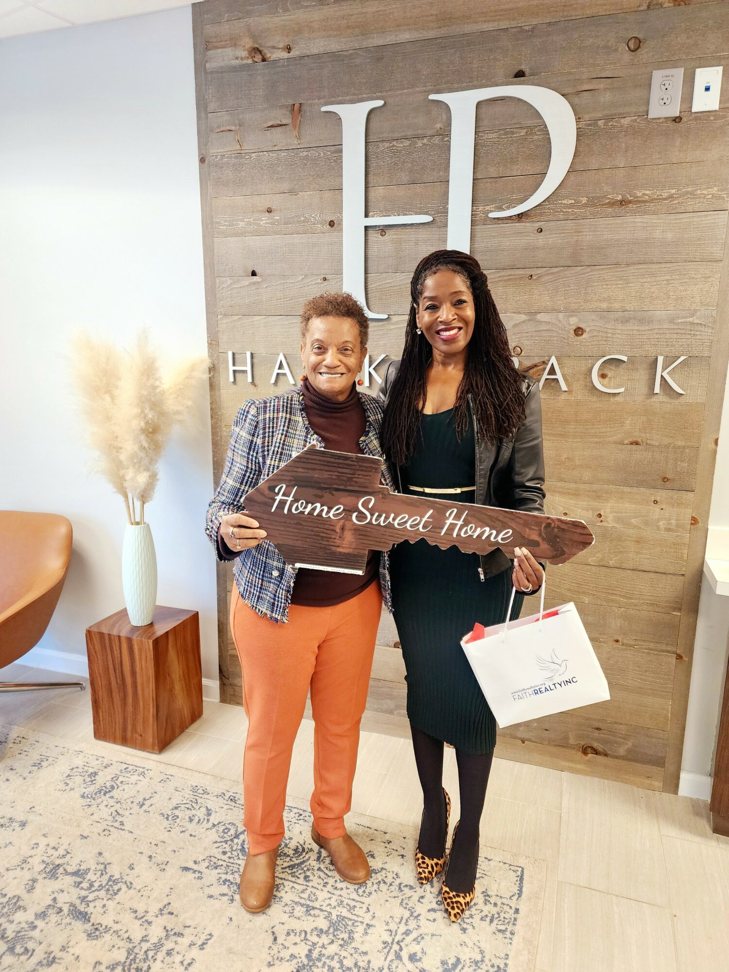 Two women standing indoors, holding a wooden sign that says 'Home Sweet Home,' smiling, with a wooden wall and decorative plant in background.