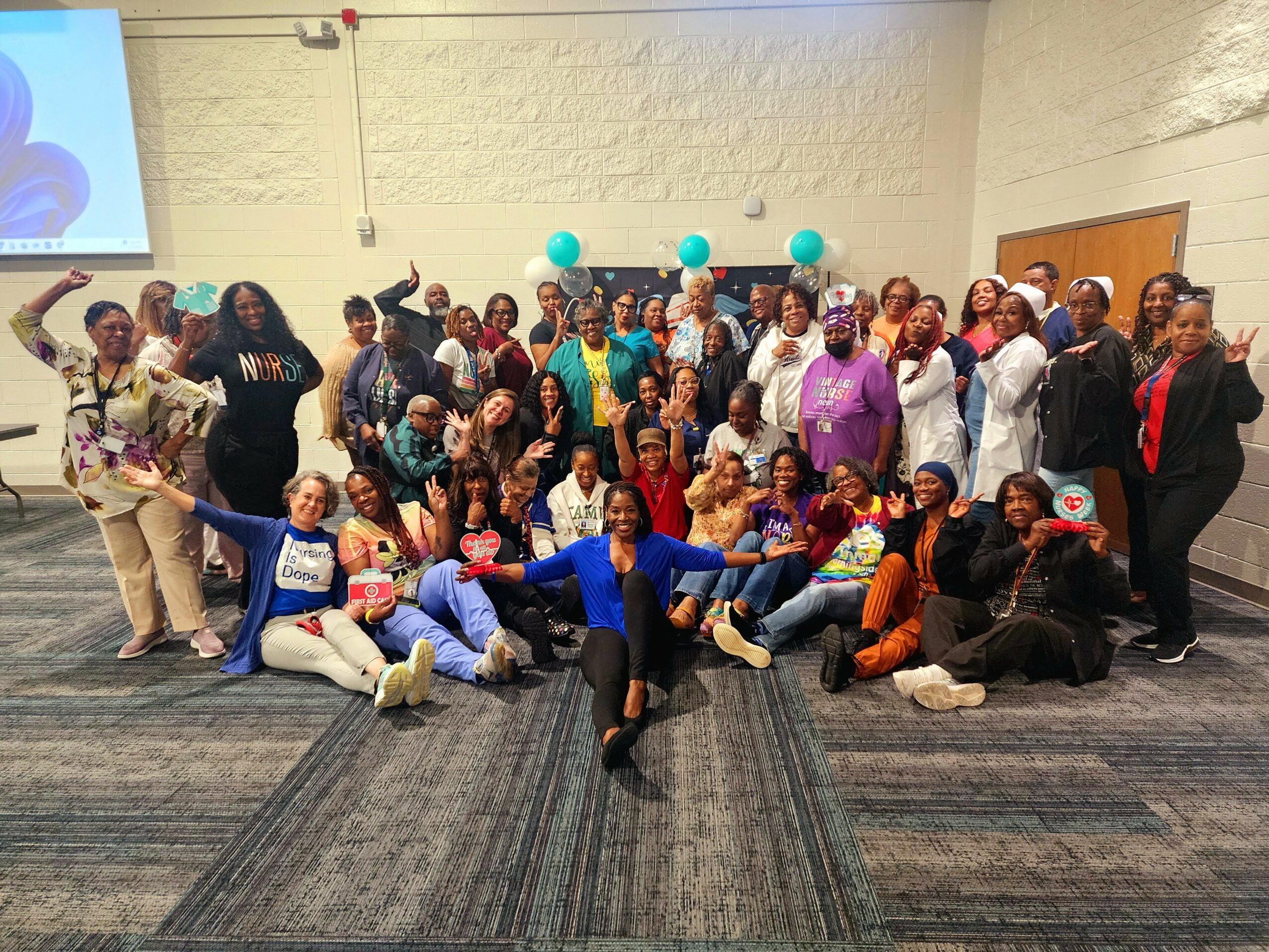 Group of diverse people posing together indoors, some sitting and some standing, with balloons in the background.