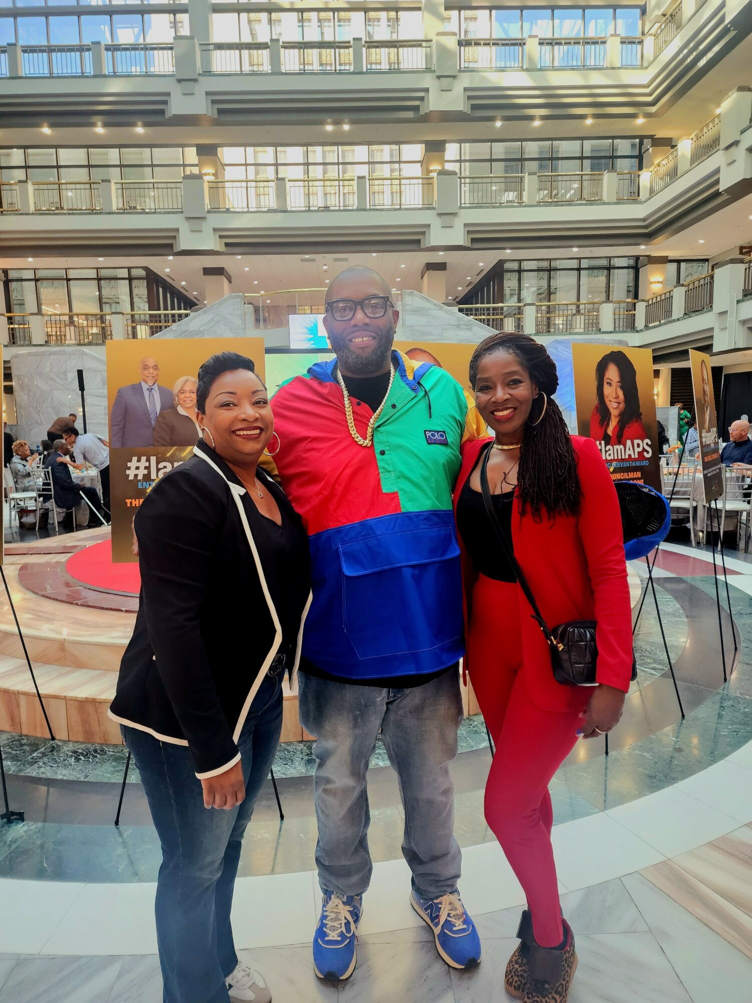 Three people standing together indoors, smiling, with a shopping mall background and promotional banners behind them.