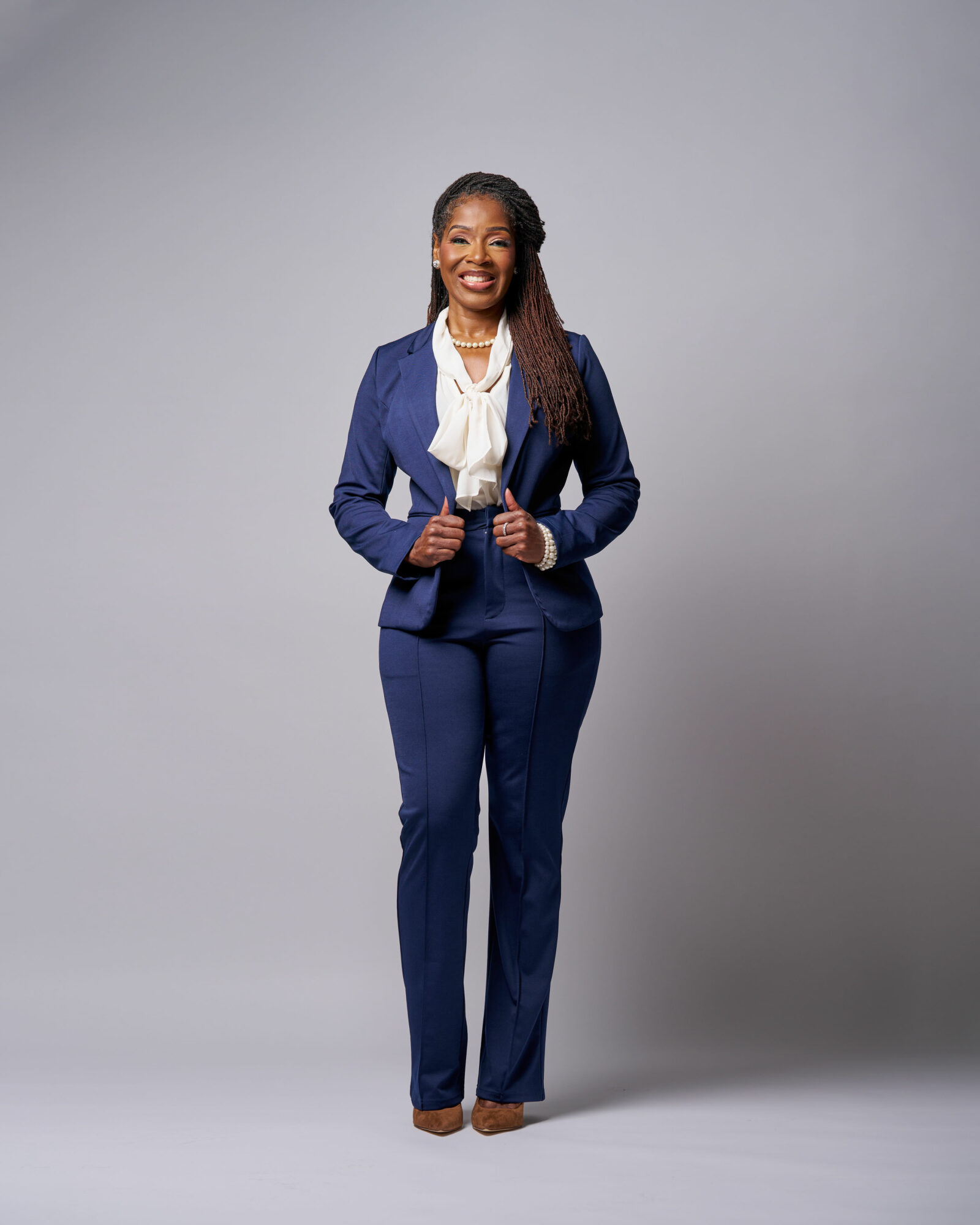 Woman in a navy blue suit with a white scarf, smiling, standing with hands on her jacket, against a plain gray background.
