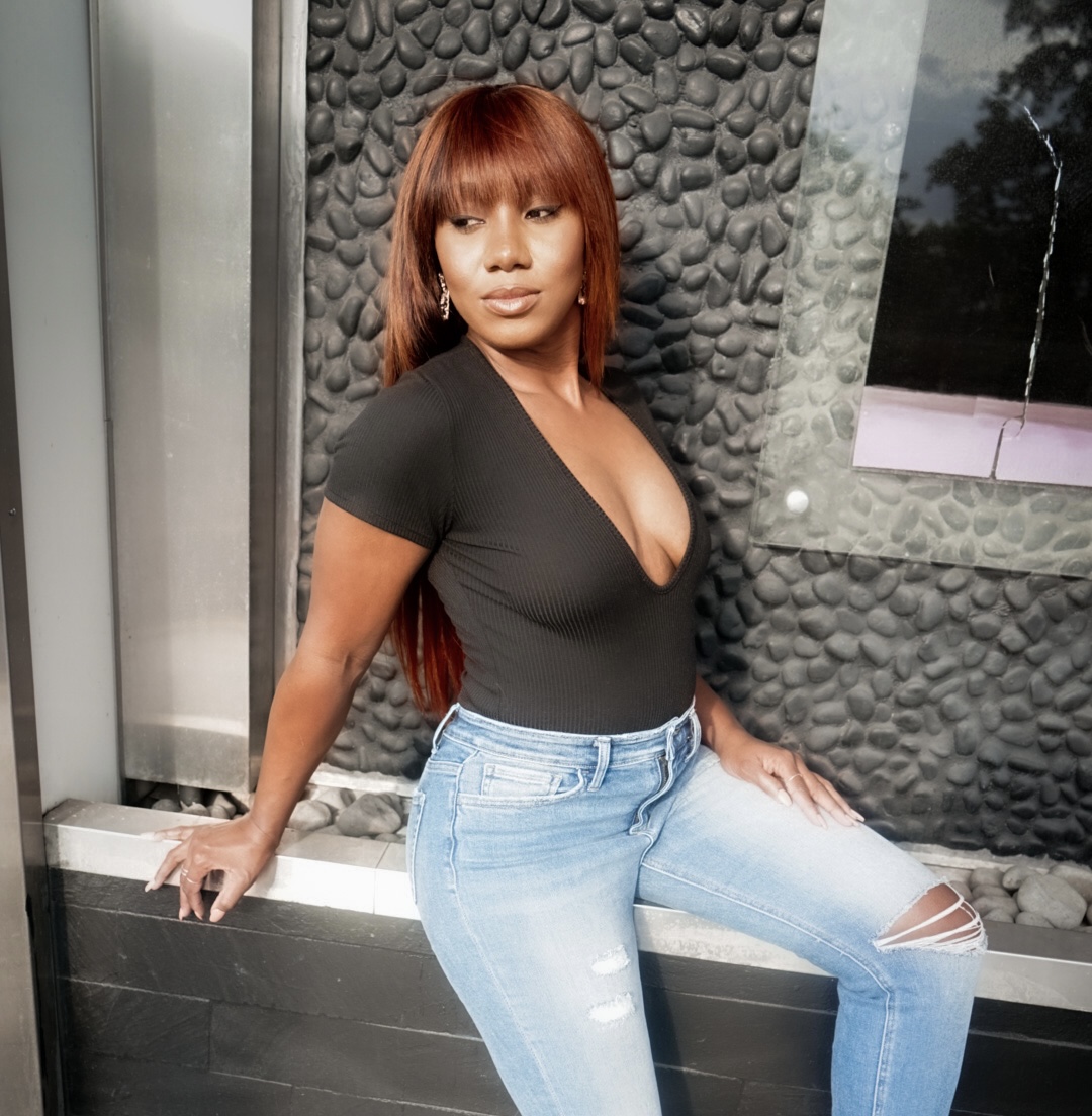 Woman with long reddish-brown hair in a black top and ripped jeans leans against a ledge outside.