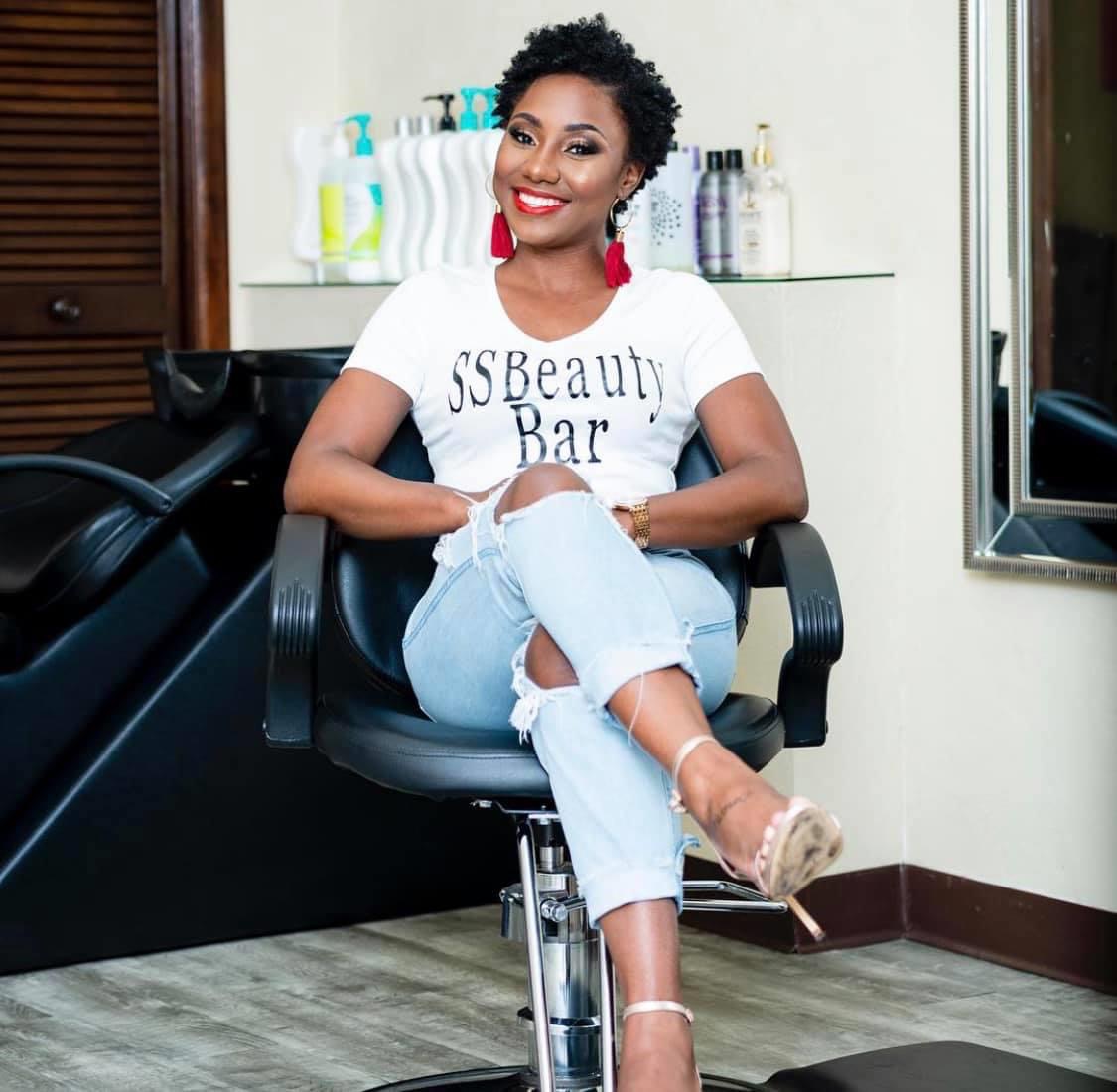 Woman with short curly hair and red earrings sitting in a salon chair, smiling, with beauty products behind her.