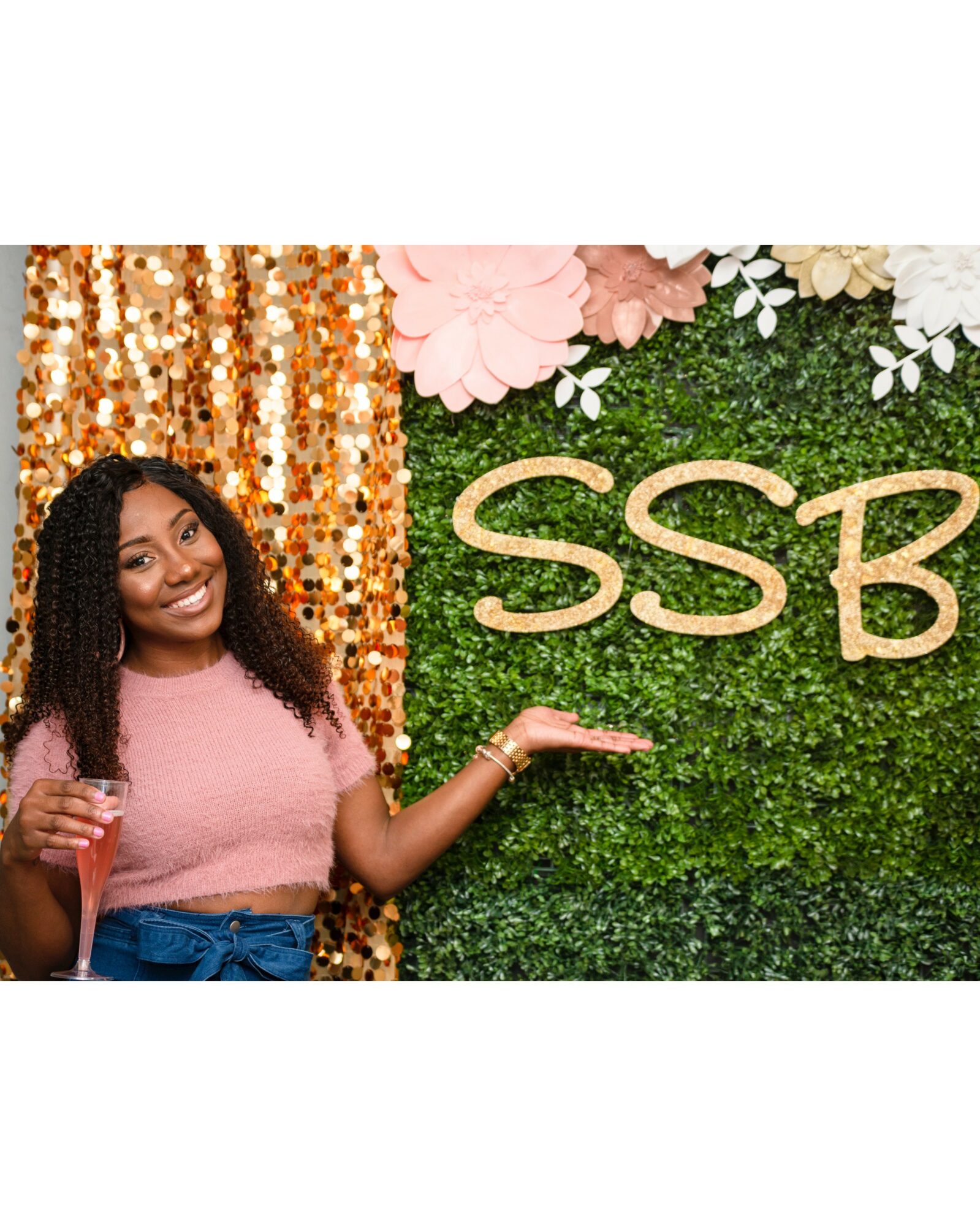 Young woman with curly hair holding a drink, standing next to a green wall with pink and white flowers and the letters 'SSB'.