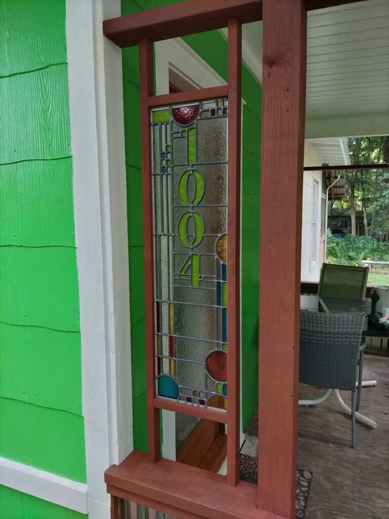 Vertical stained glass panel with the year 2004, framed by a wooden porch with outdoor furniture visible in the background.