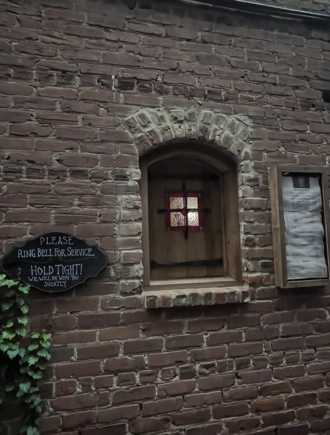 Brick wall with small window, sign, and a framed object, with greenery at the bottom left corner.
