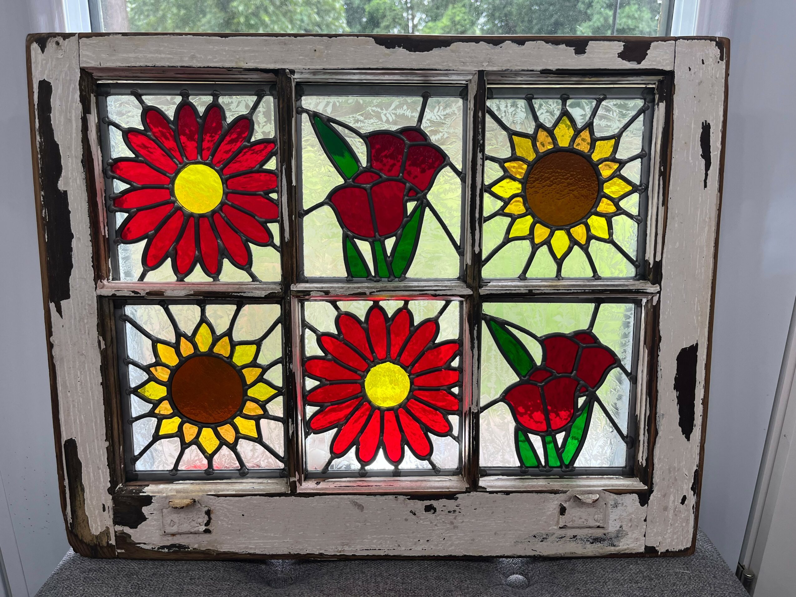 Colorful stained glass window with flowers, including sunflowers and roses, in a distressed white wooden frame.