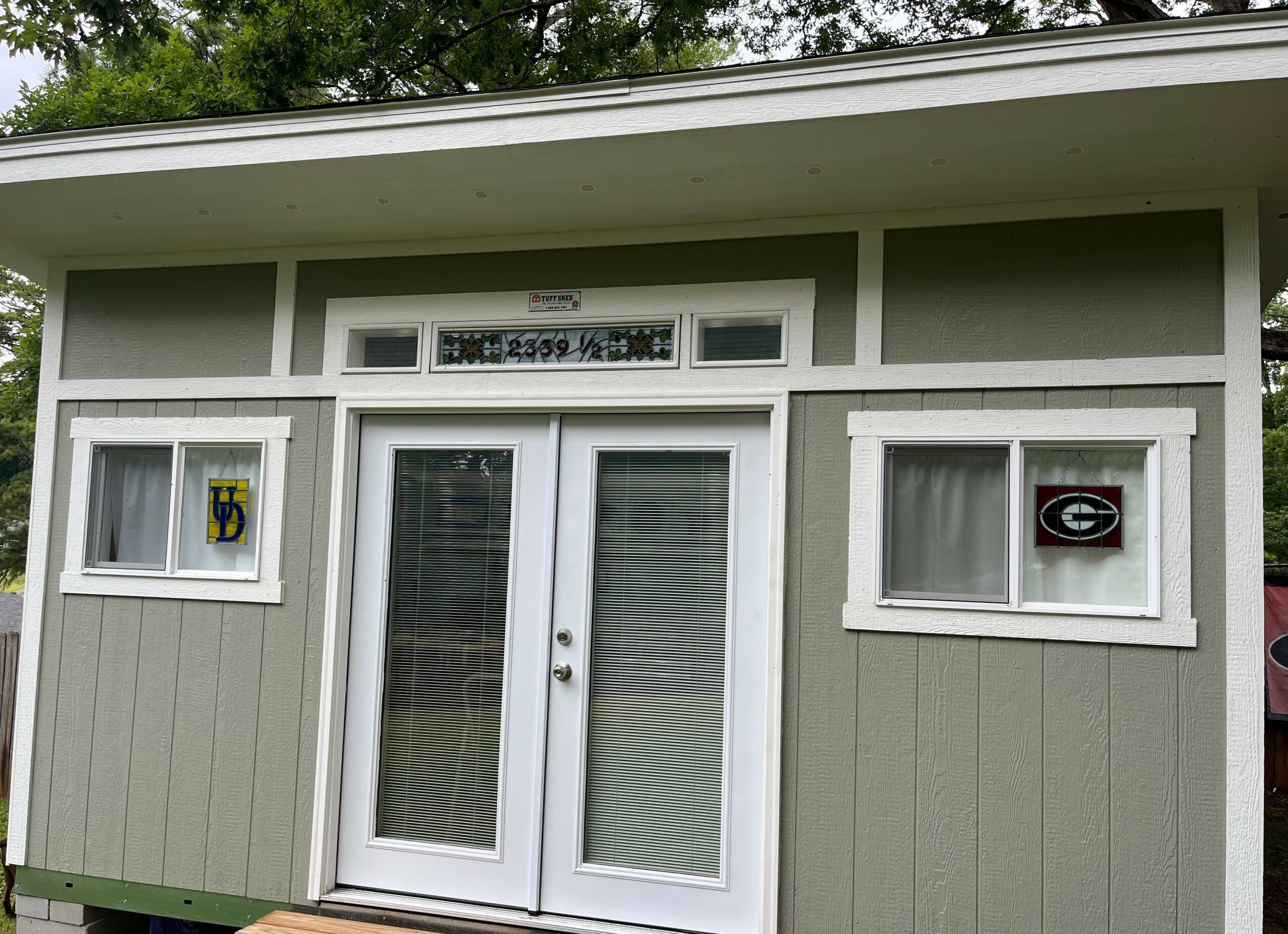 Small building with double glass doors and two windows, surrounded by green trees, painted in light green with white trim.