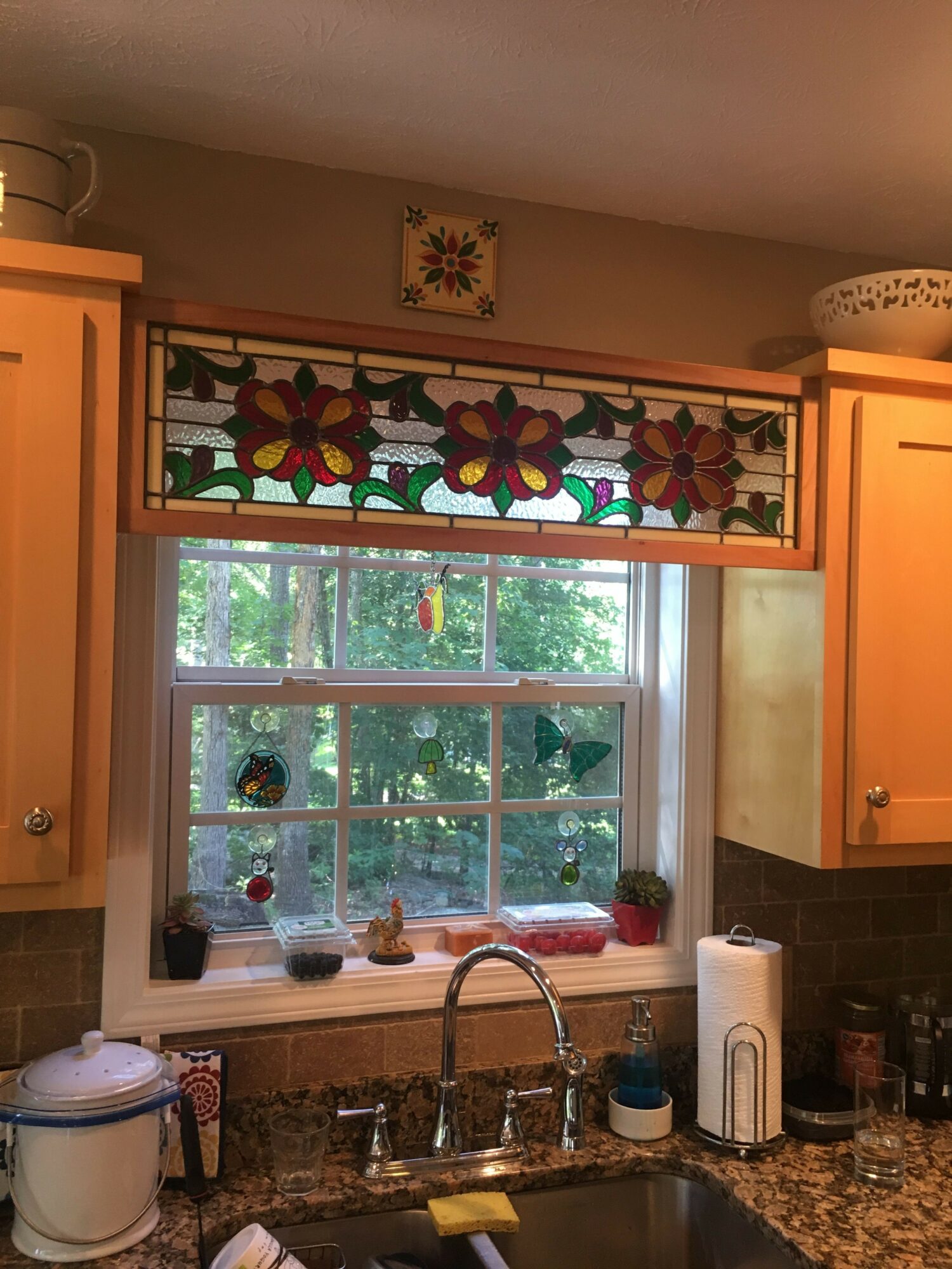 Kitchen window with stained glass floral design, view of trees outside, sink with faucet, and kitchen items on counter.