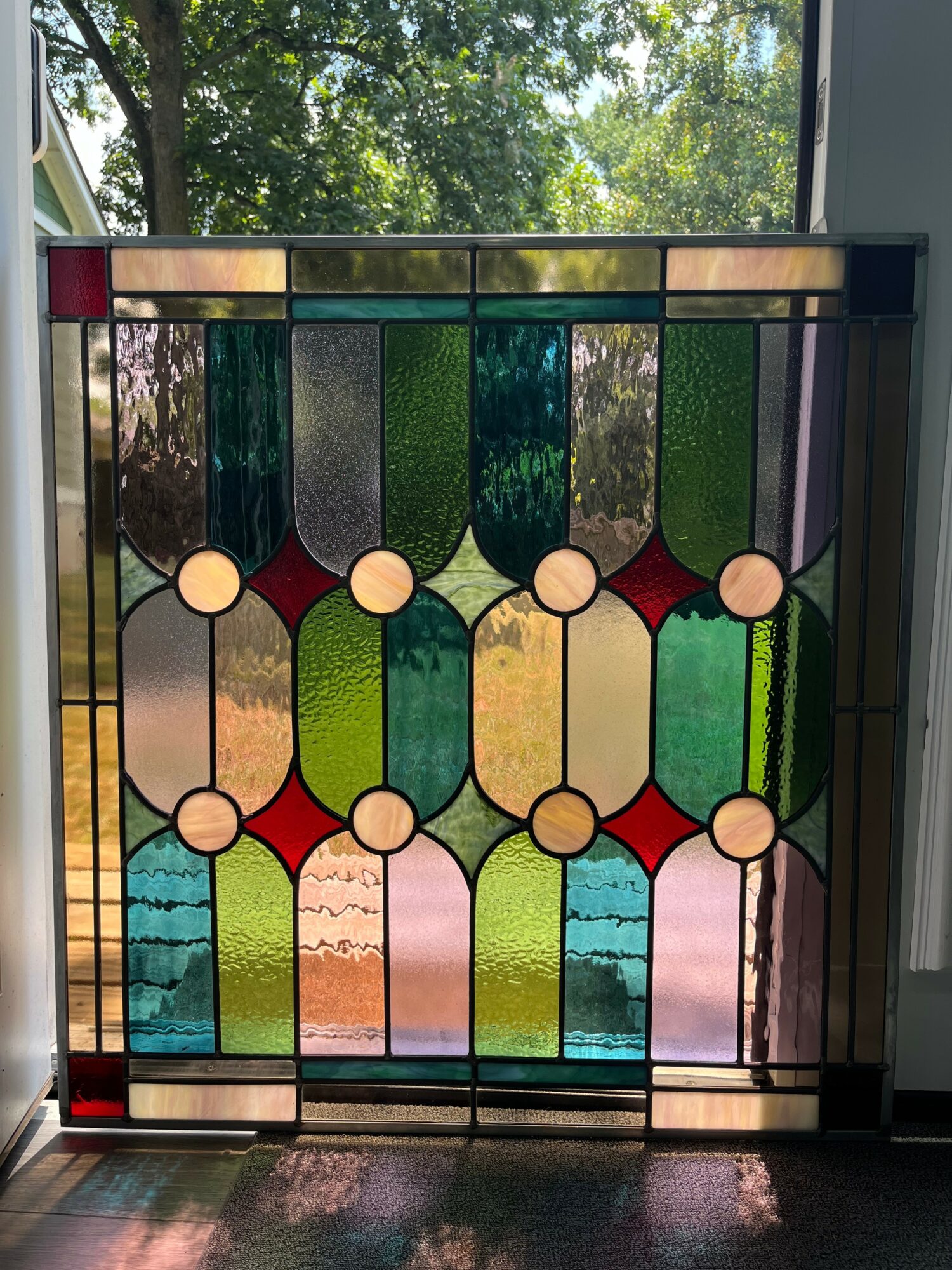 Colorful stained glass window with geometric patterns and circular accents, sunlight shining through, trees visible outside.