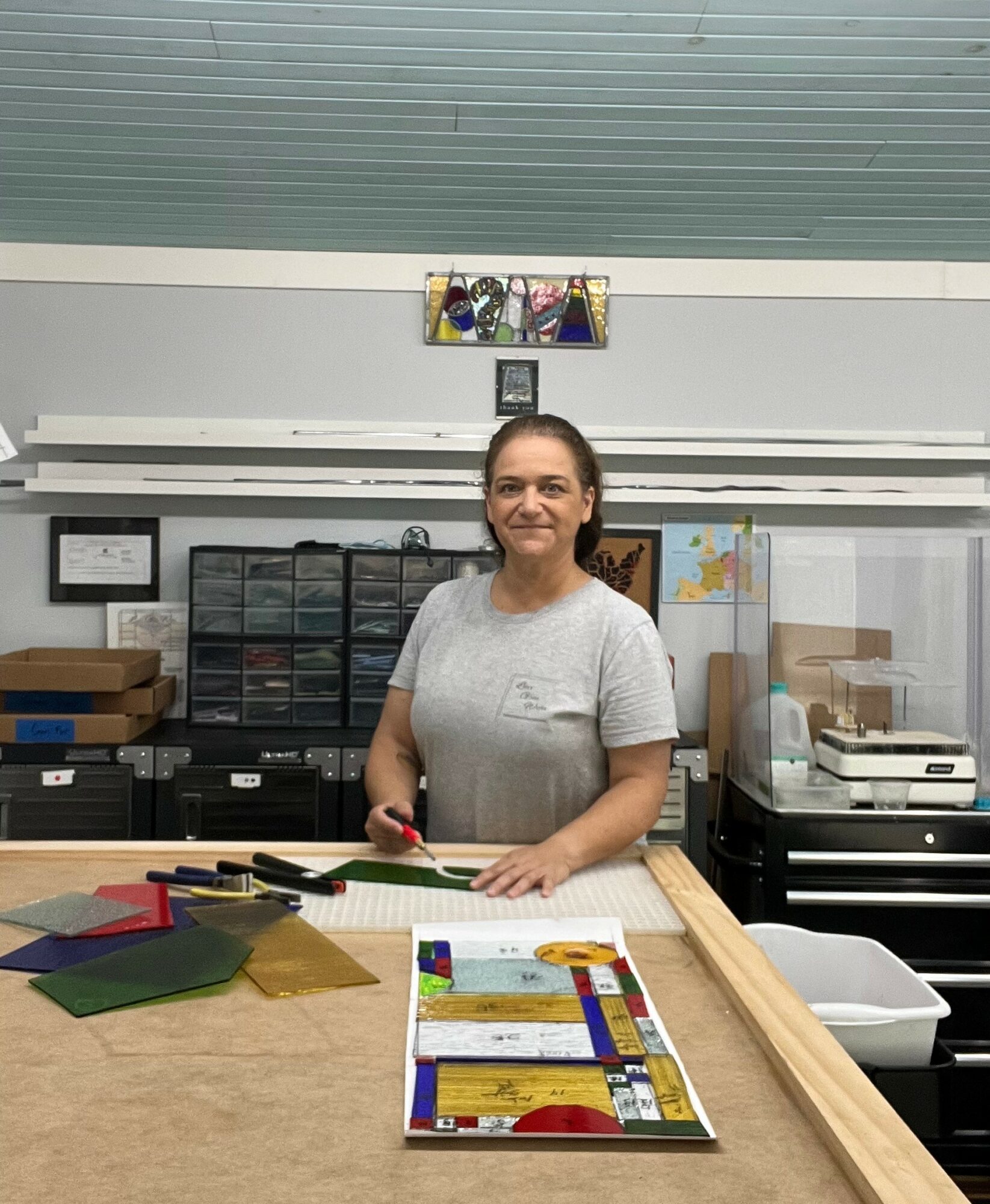 Woman standing behind a worktable with craft supplies, smiling, in a workshop or studio setting.