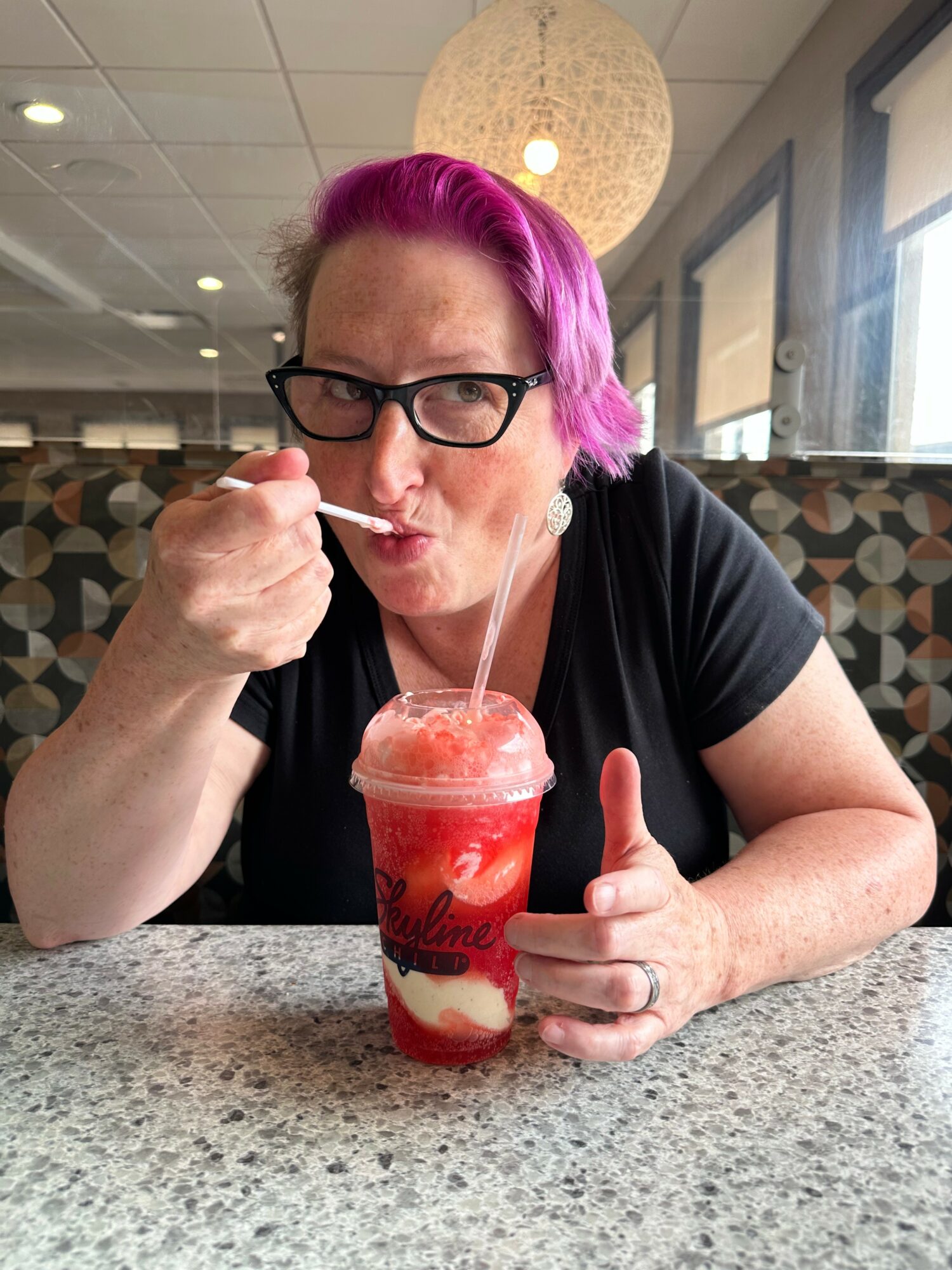 Person with pink hair and glasses eating a dessert in a glass jar at a table.