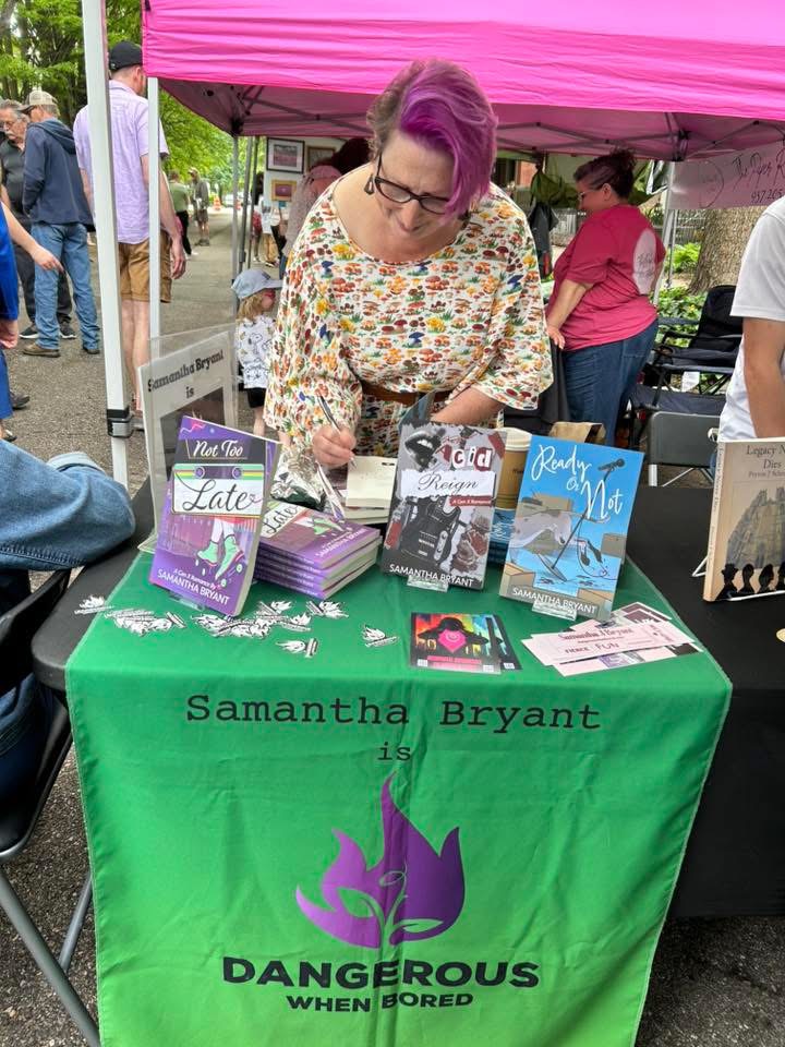 Woman with purple hair and glasses at a book signing table with books and a green tablecloth, outdoors, pink canopy overhead.