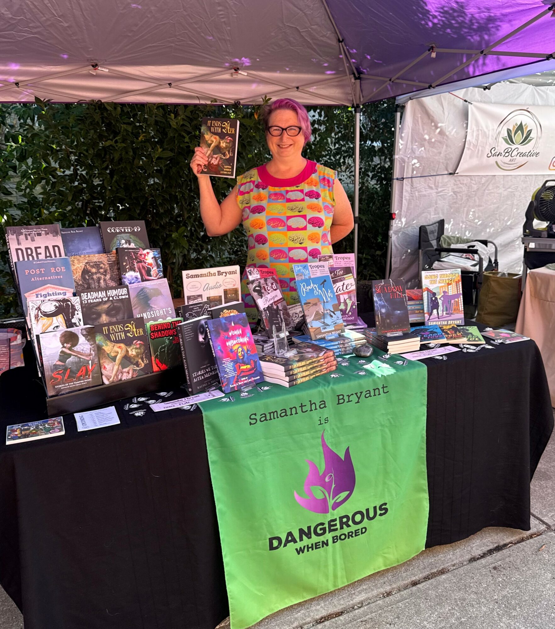 Woman standing behind a book table under a canopy, holding a book, with books displayed on the table and a green banner reading 'Samantha Bryant DANGEROUS WHEN BORED'.