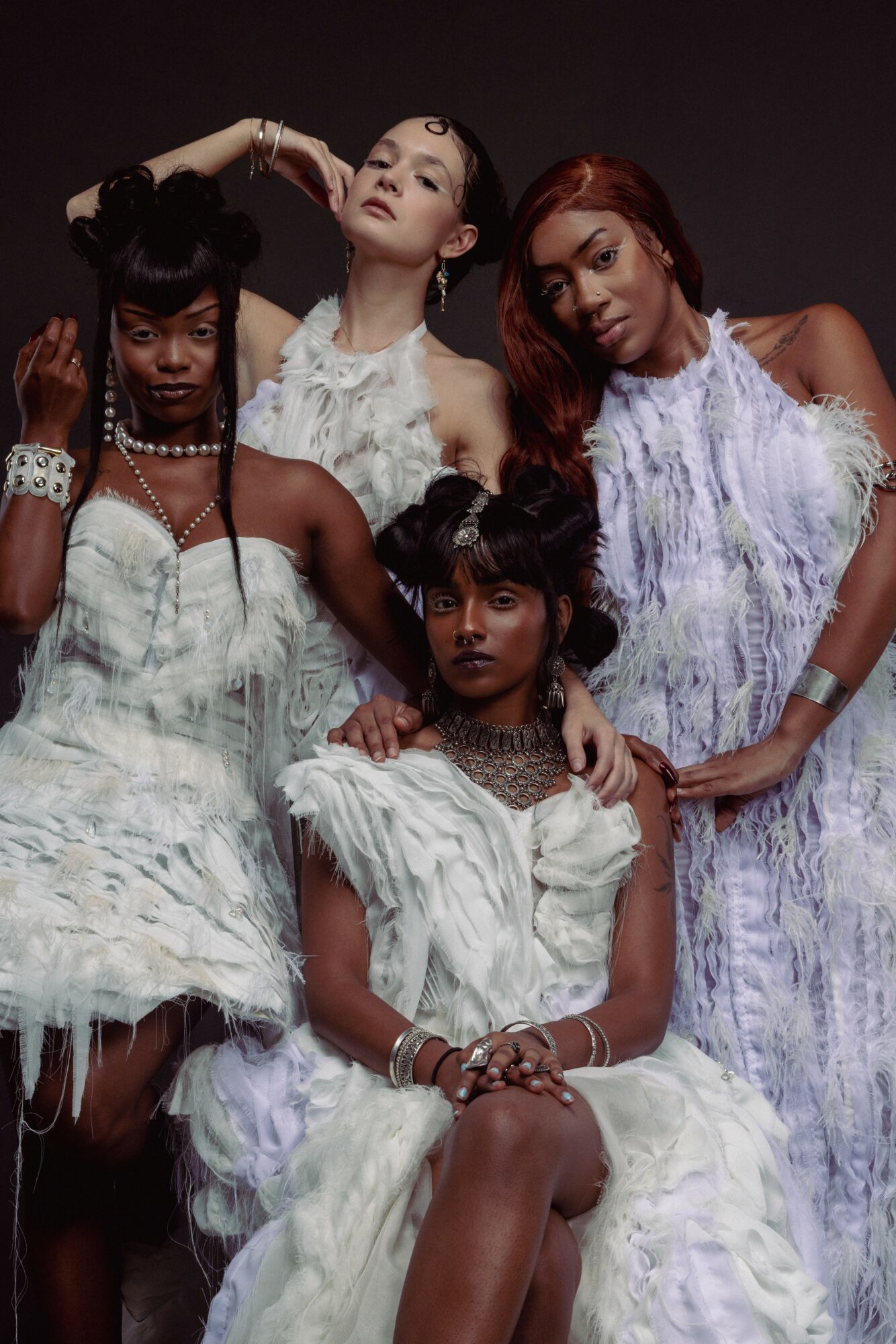 Four women in elegant dresses pose together against a dark background, with serious expressions and stylish hairstyles.