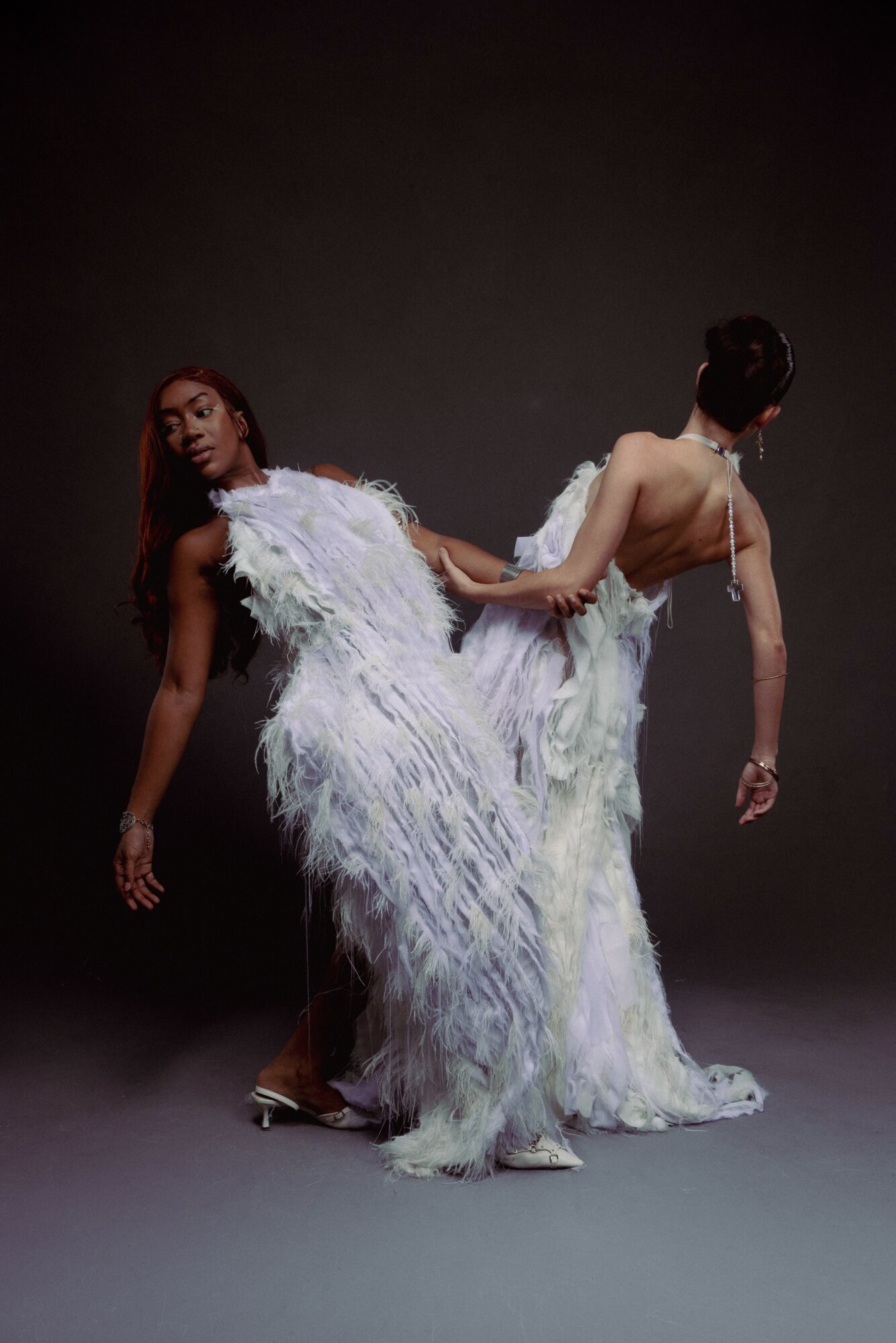 Two women in white feathered dresses pose against a dark background, one facing forward and the other turned away.