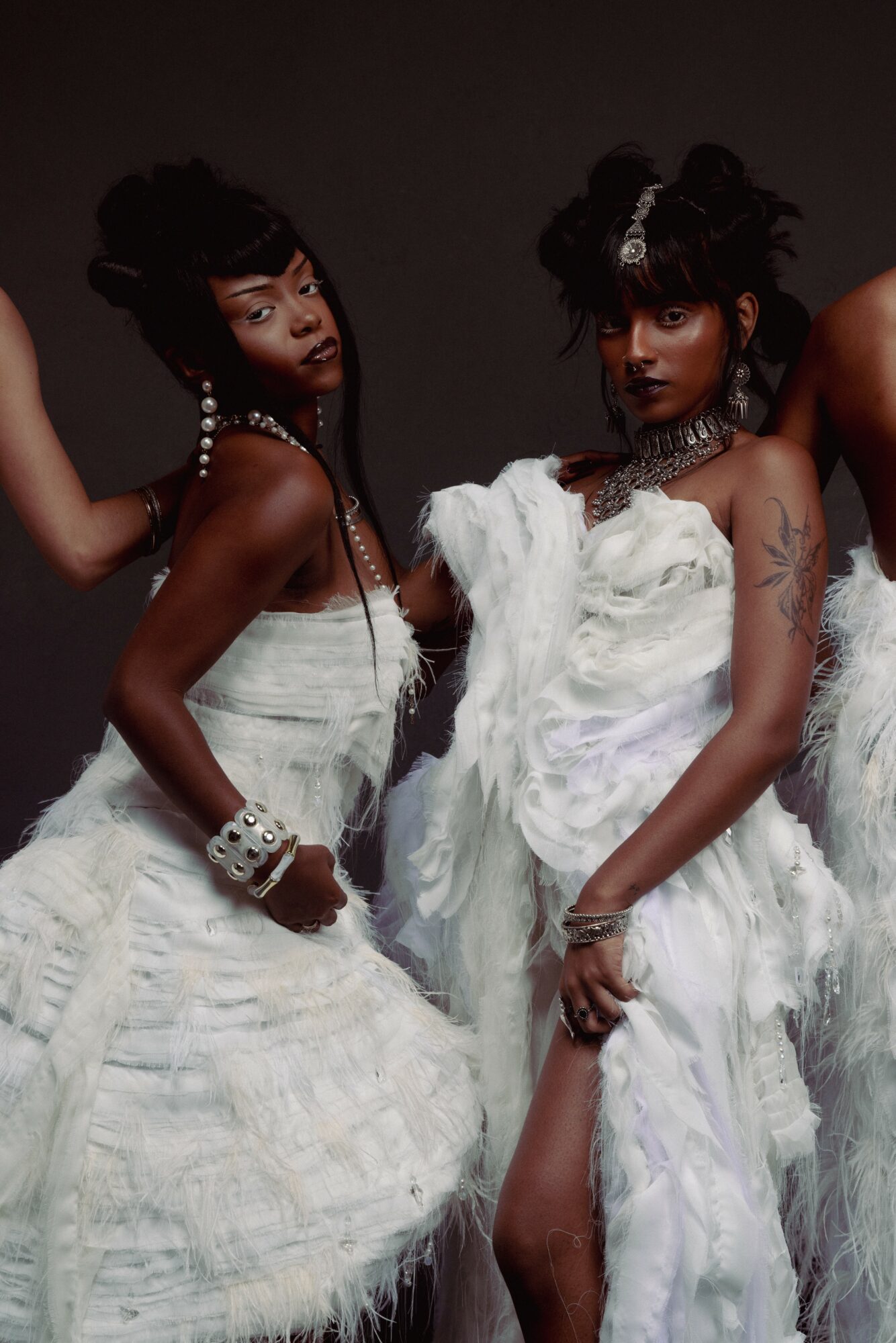 Two women in white dresses with jewelry pose against a dark background, one with a hand on her hip and the other holding her dress.