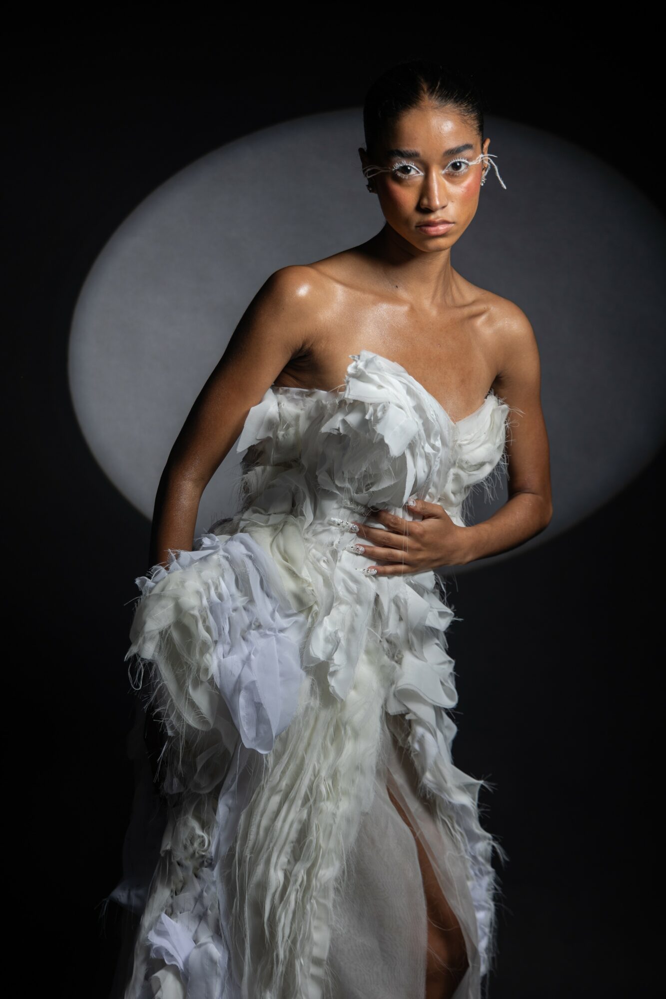 Woman in a textured, flowing white dress with glasses, standing against a dark background with a circular light behind her.