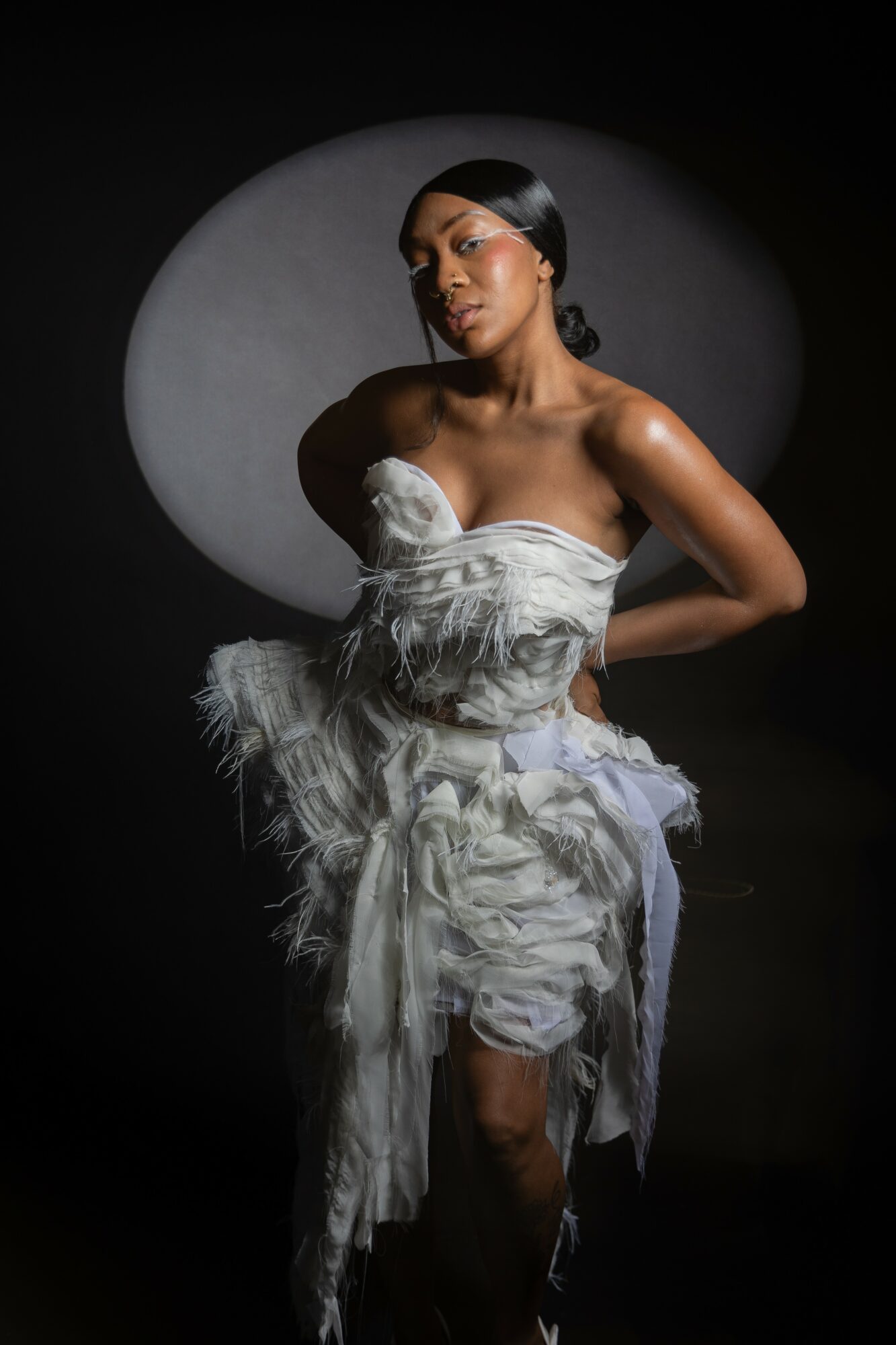 Woman in a strapless, ruffled white dress poses against a dark background with a circular light behind her.