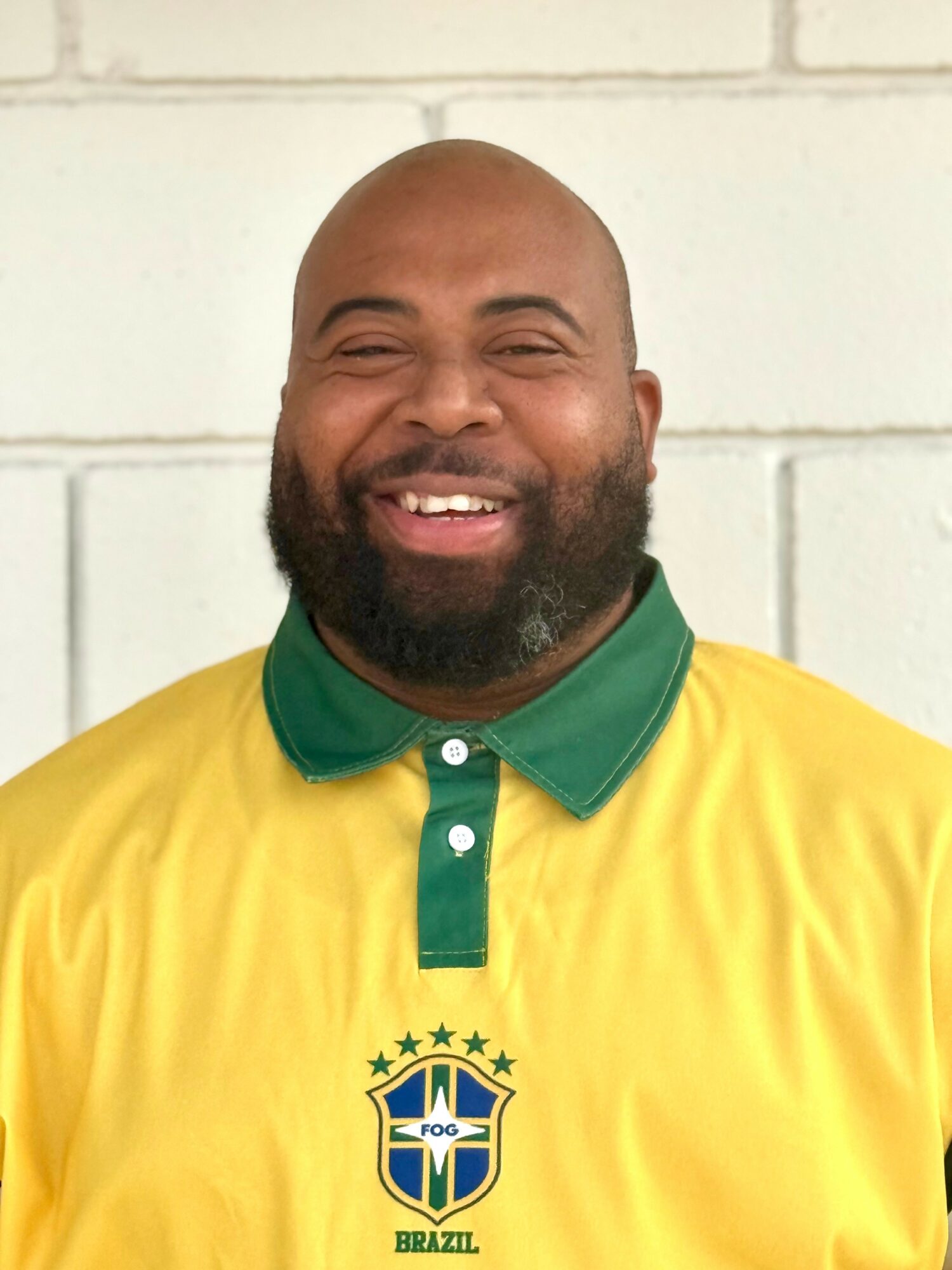 Smiling man with a beard wearing a yellow and green Brazil soccer jersey, standing against a white brick wall.
