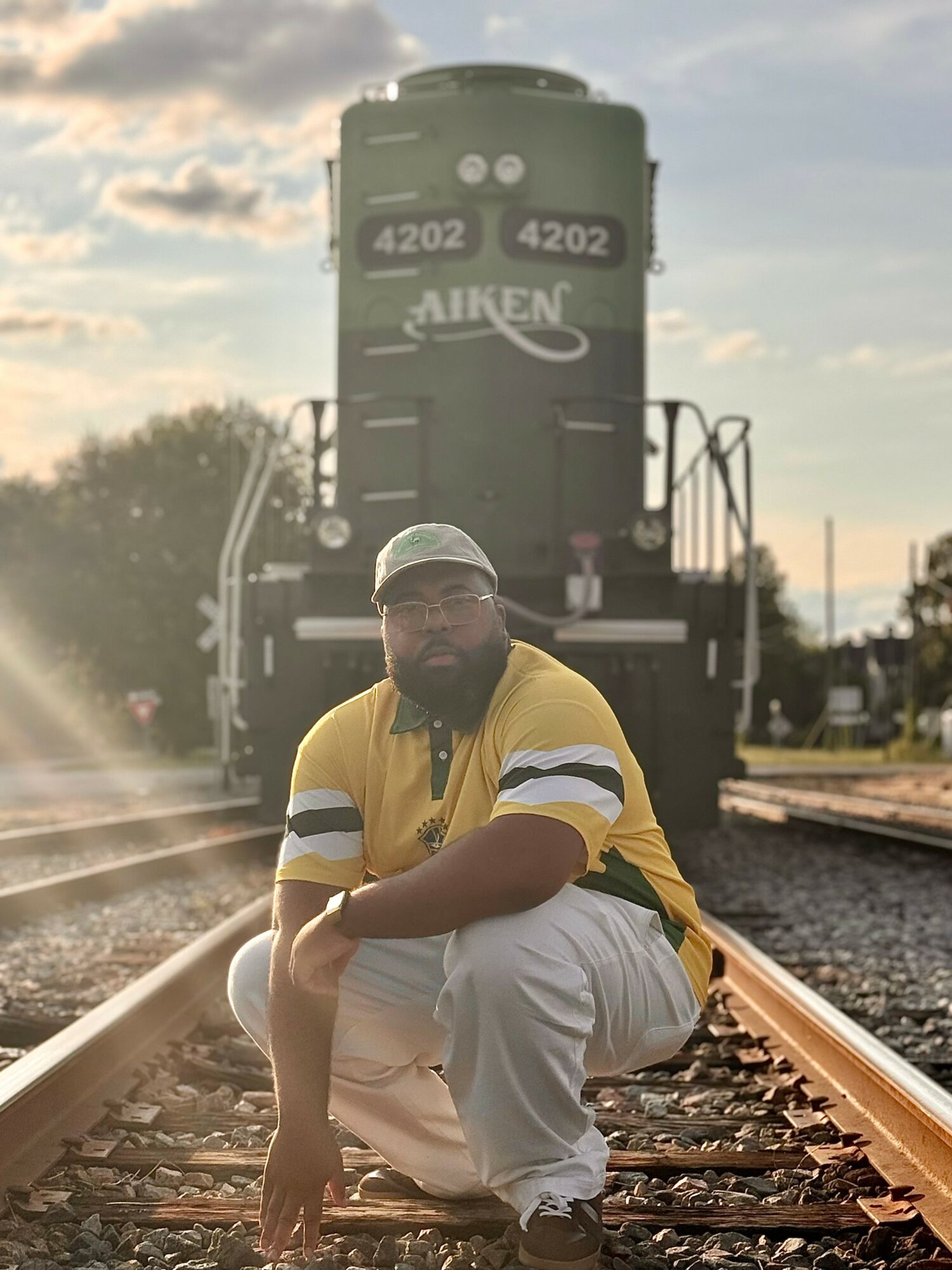 Man crouching on railroad tracks with a large train behind him, sunset sky, wearing a yellow and black shirt, white pants.