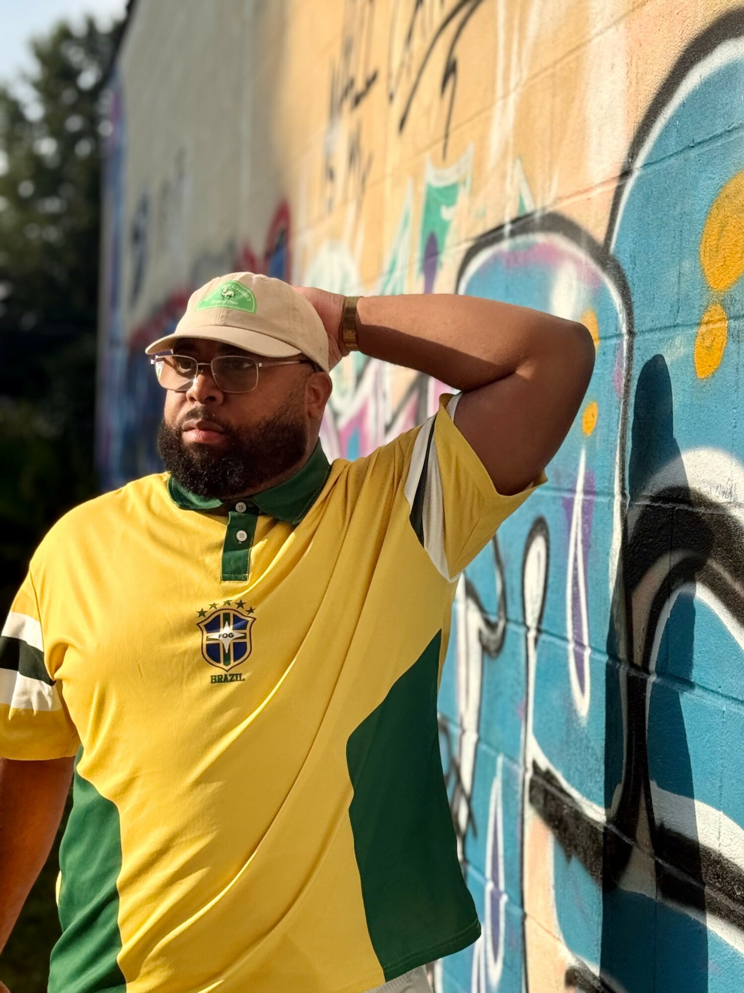 Man with glasses and beard wearing a yellow and green sports jersey, standing next to a colorful graffiti wall.