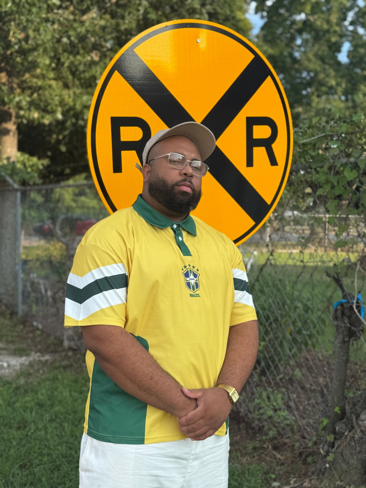 Man with glasses and beard wearing a yellow and green sports jersey, standing outdoors in front of a large yellow and black road sign.
