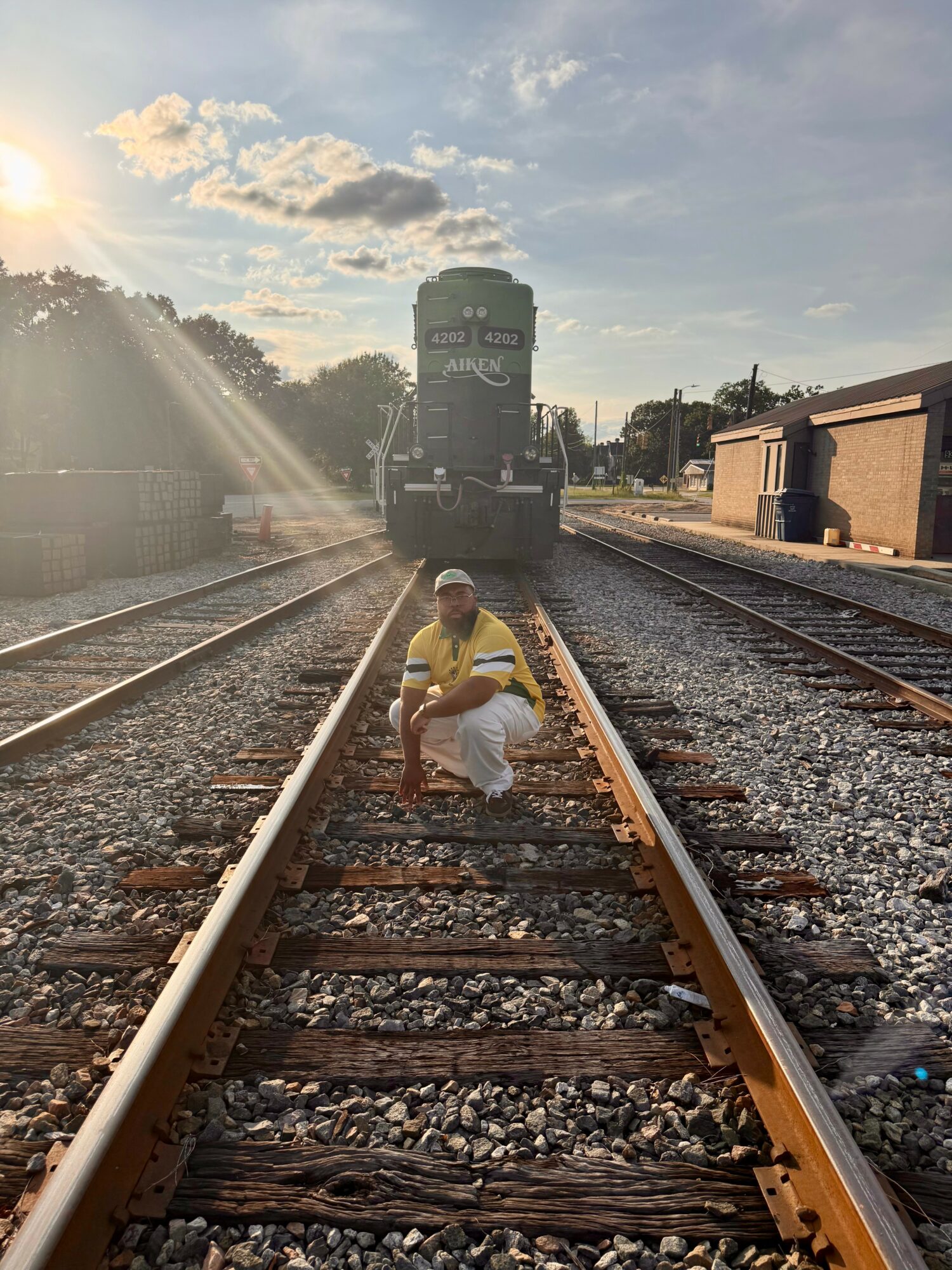 Person crouching on railway tracks with train approaching in the background, sunlight shining from the left, sky partly cloudy.