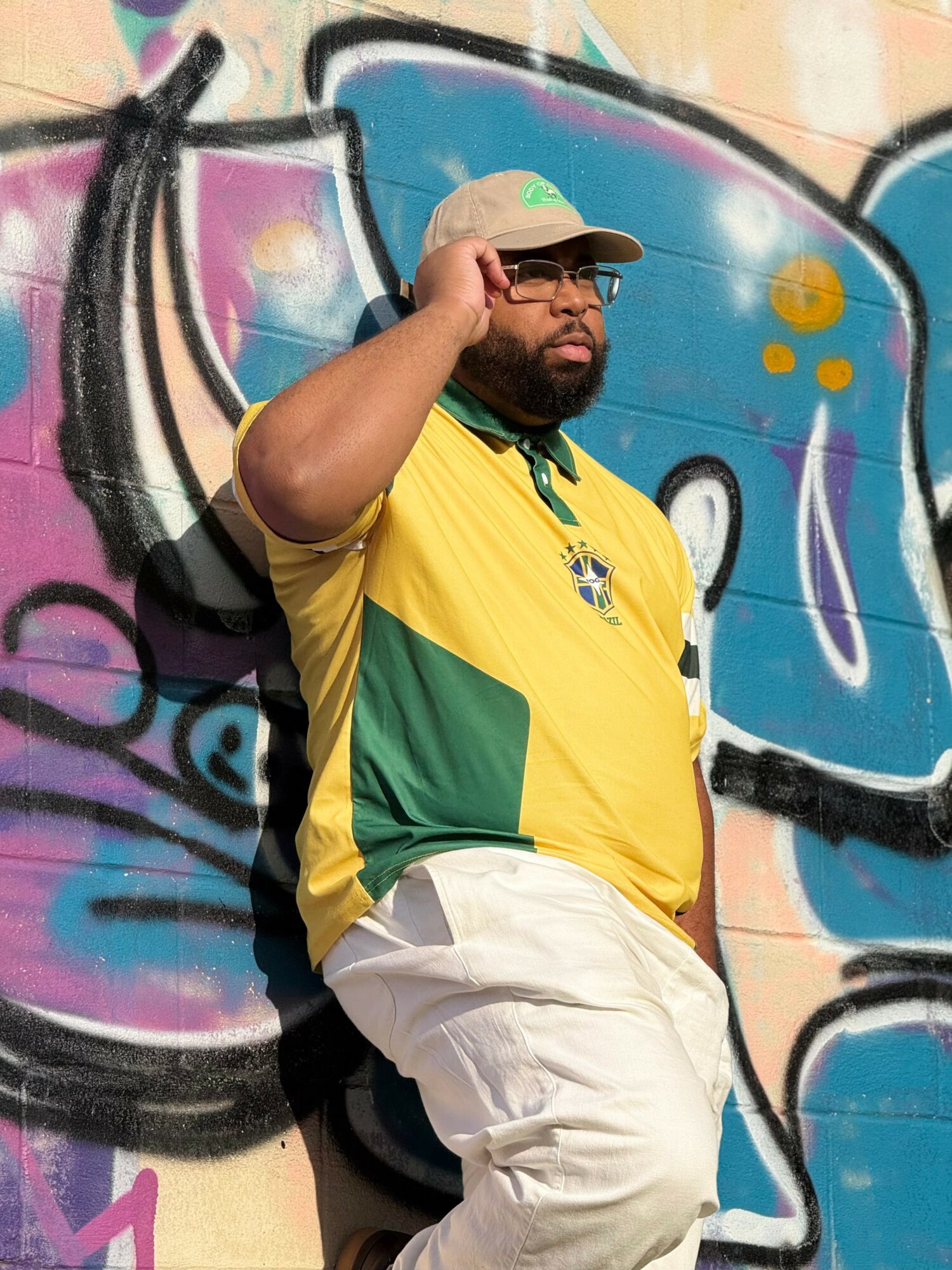 Man with glasses and beard wearing a yellow and green shirt, beige pants, and a cap, standing against a colorful graffiti wall.