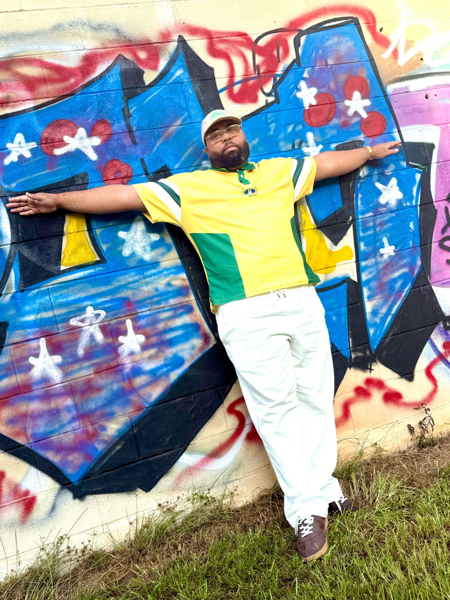 Person standing with arms outstretched in front of colorful graffiti wall, wearing a yellow and green shirt and white pants.