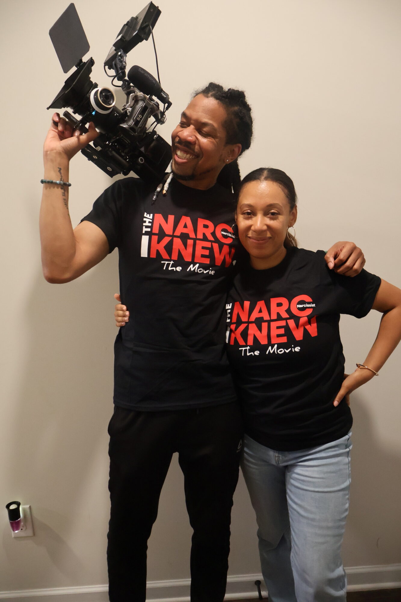 Two smiling people wearing black T-shirts with red text, one holding a professional video camera, standing against a plain wall.