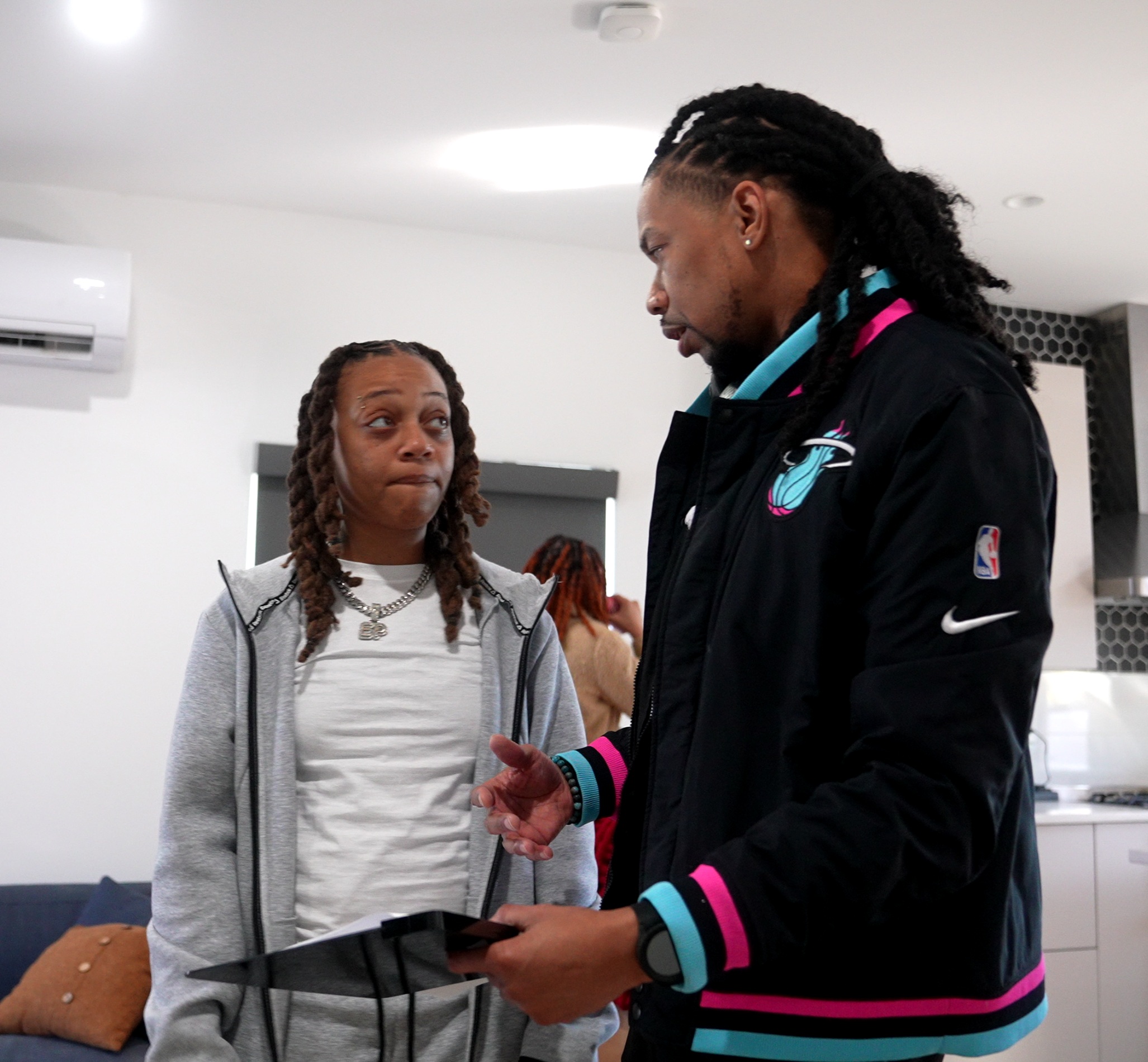 Two people talking indoors, one holding a tablet, in a modern room with white walls and an air conditioner.