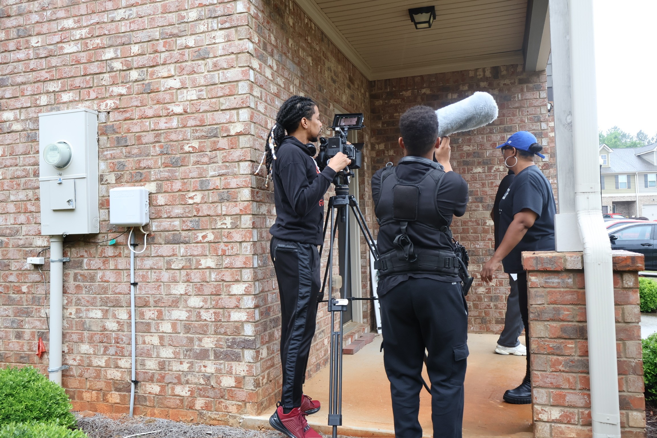 Three people standing outside a brick building, one with a camera on a tripod, another holding a boom microphone, and a third person in the background. Word count: 20, confidence: 0.9