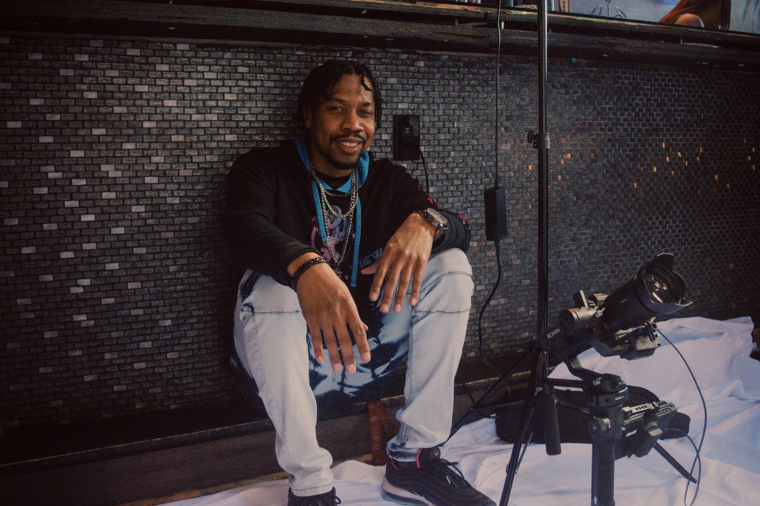 Person with curly hair and beard sitting on the floor, smiling, in front of a dark tiled wall, with camera equipment nearby.