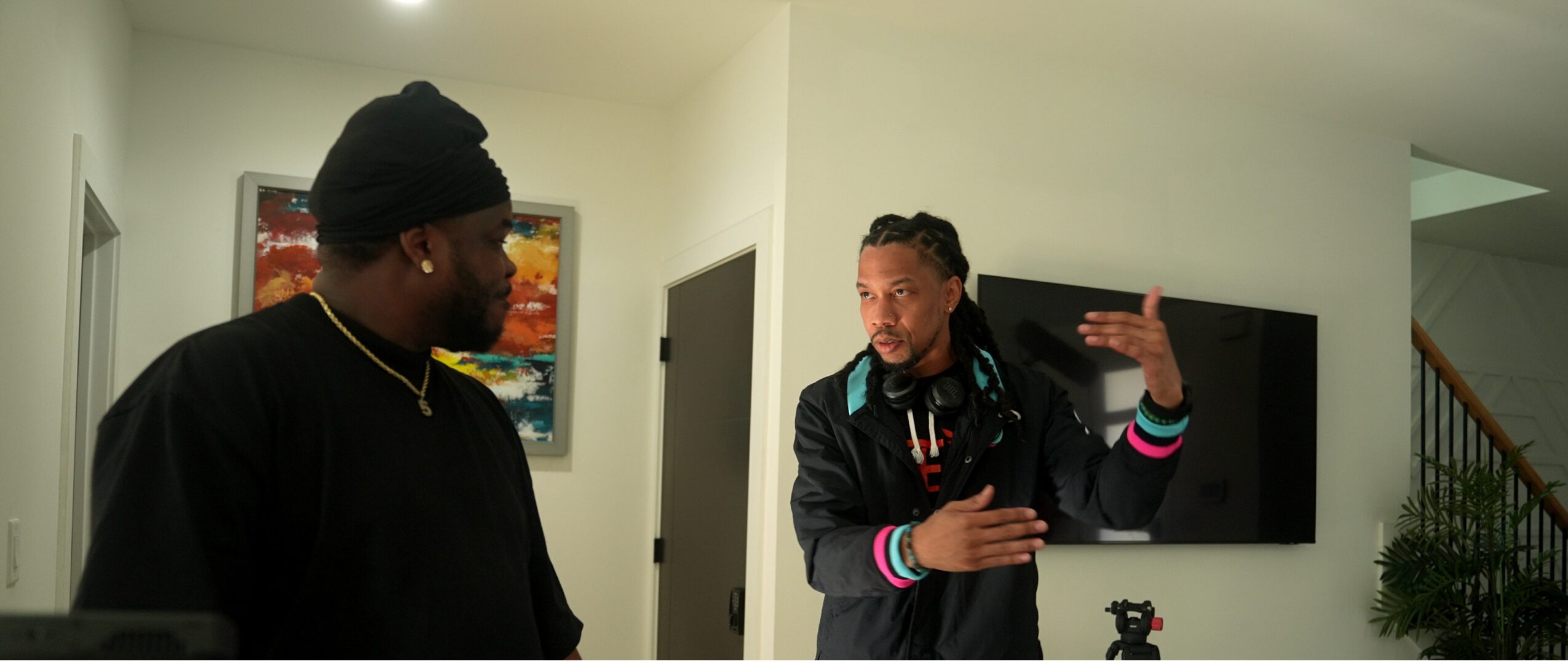 Two people engaged in conversation indoors, one gesturing with hand, the other listening, with a television and staircase in background.