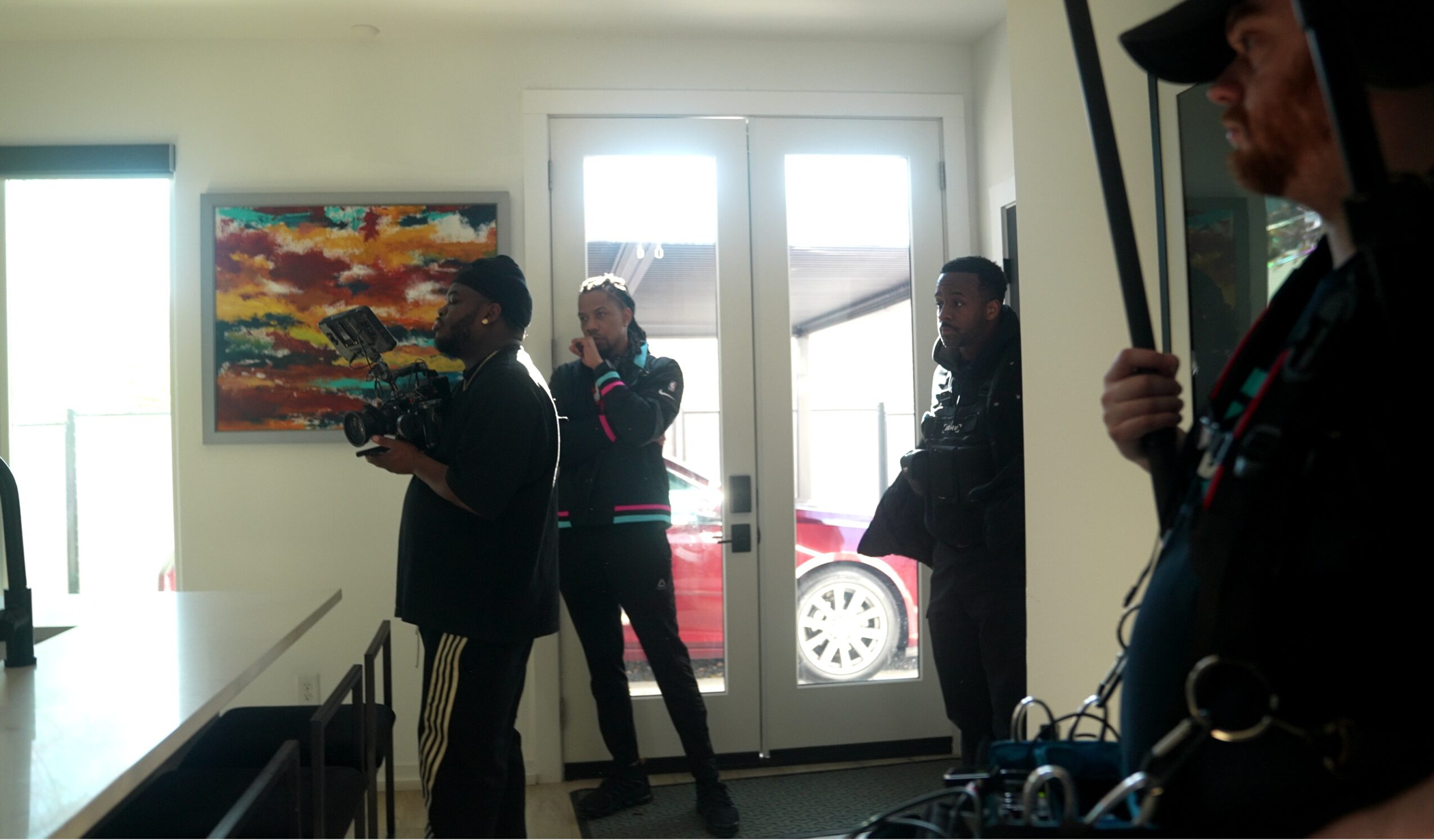 Four people stand inside a room near a door with a window, one holding a drone controller, others holding objects, a colorful painting on the wall.