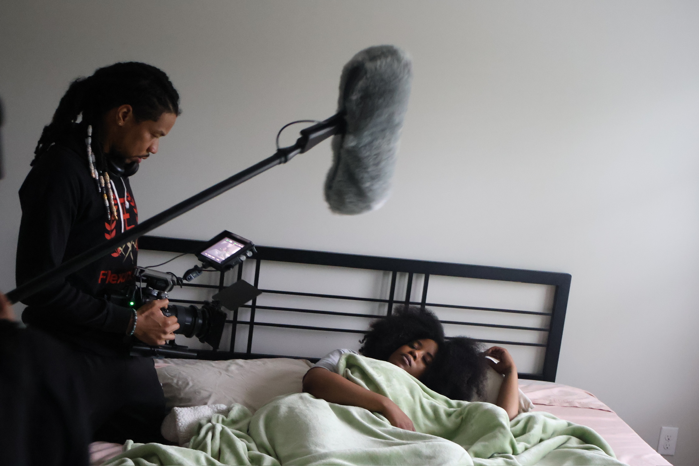 Person lying in bed with a blanket, another person standing nearby, recording with a camera and microphone, plain wall background.