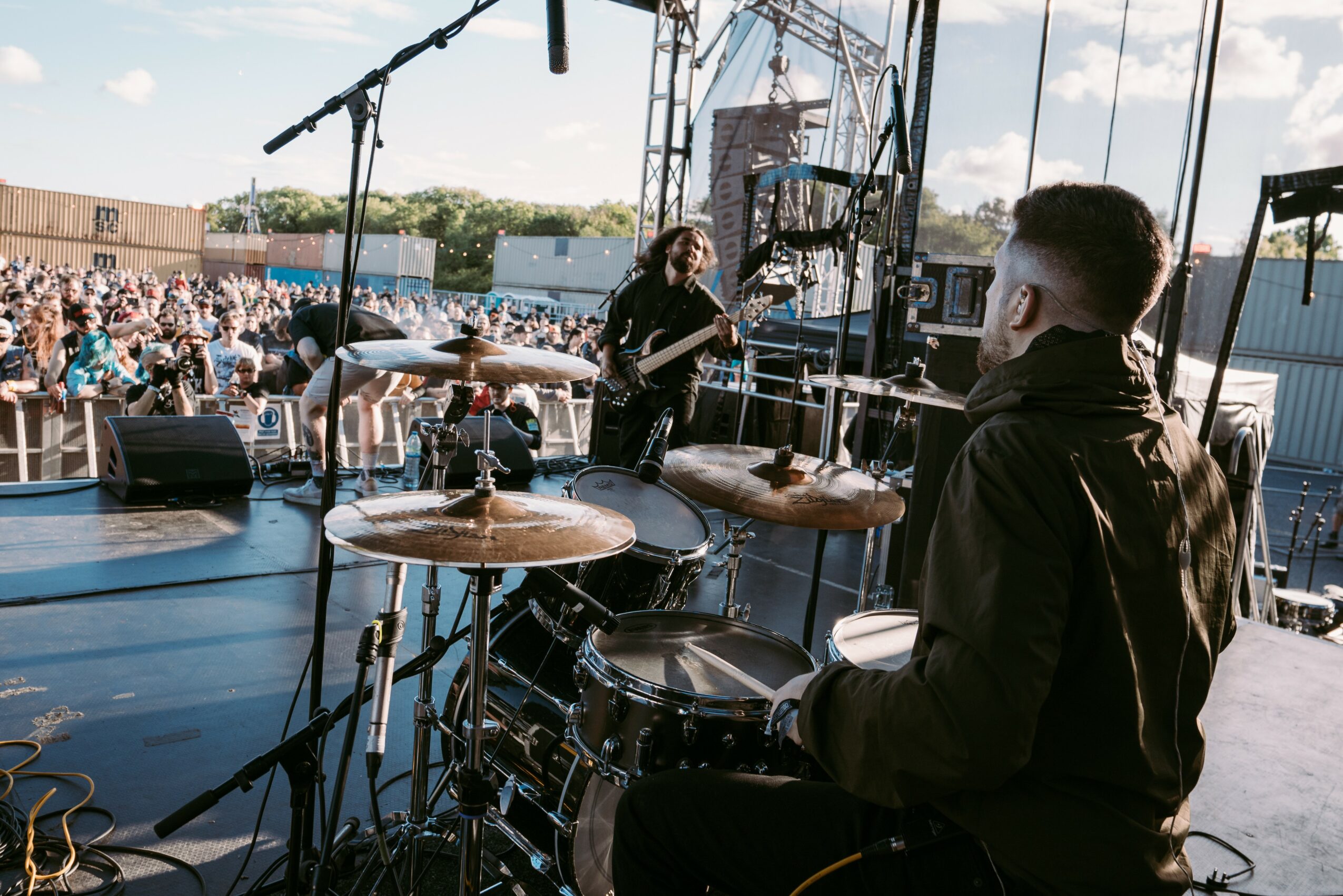 Musicians perform on an outdoor stage with a crowd in the background, drums and a microphone visible.