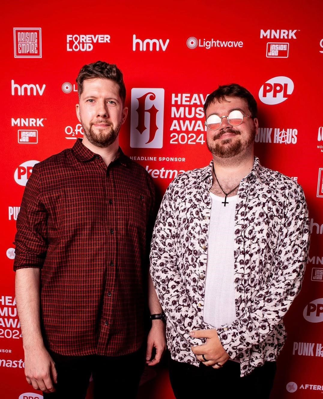 Two men standing in front of a red backdrop with event logos, one wearing a red checkered shirt, the other in a patterned jacket.