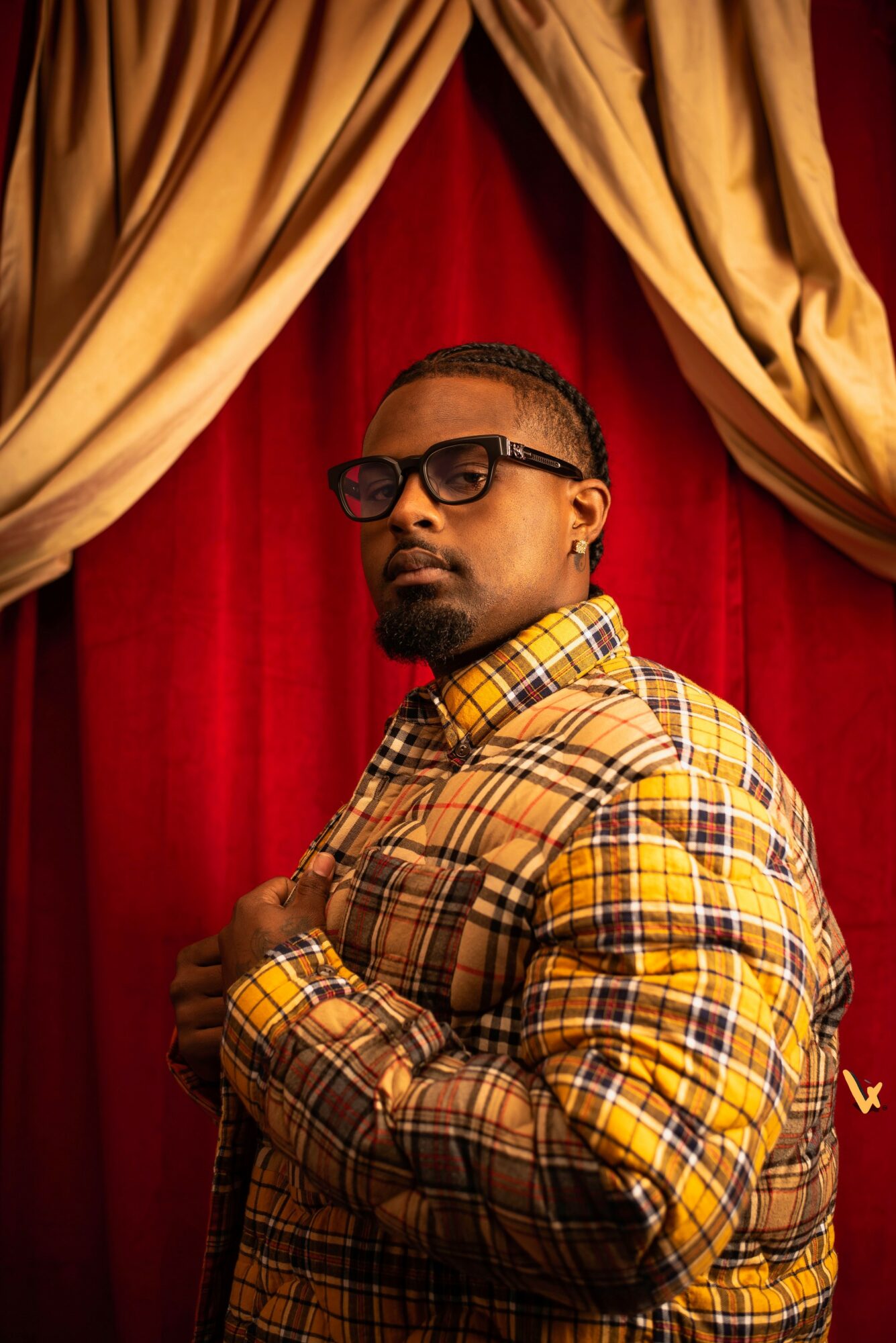 Man with glasses in plaid shirt stands in front of red curtain with gold drapes, looking at camera.