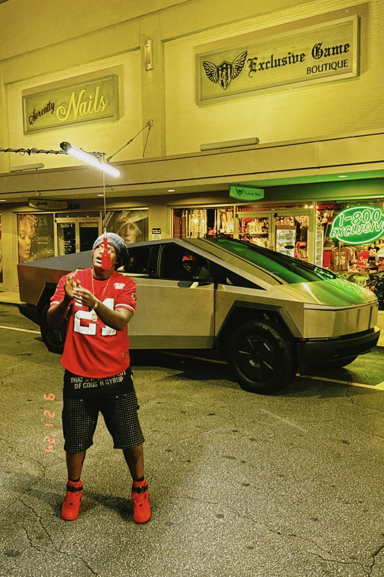 Person in red shirt and shorts standing in front of a futuristic car outside a store at night.
