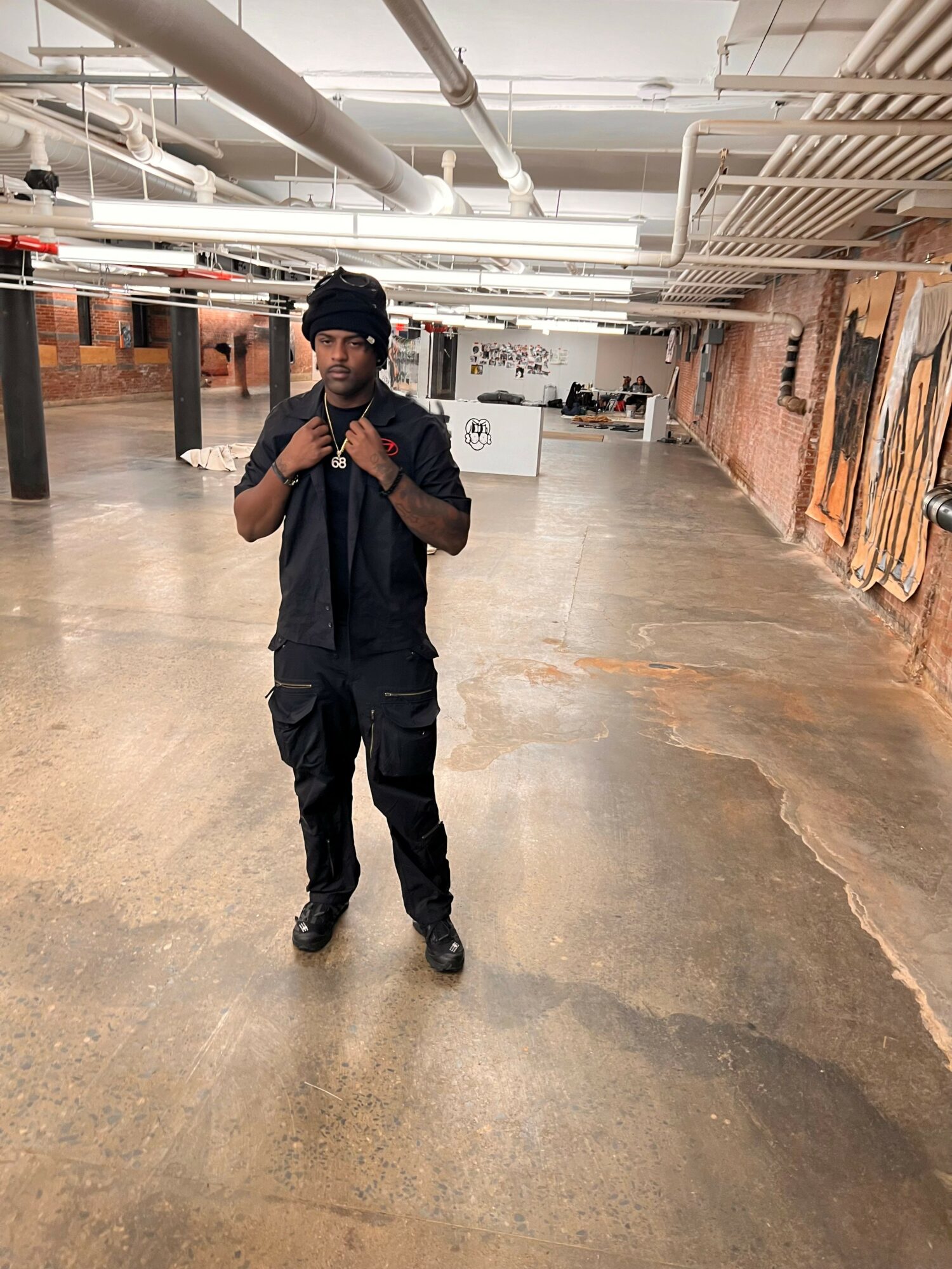 Person standing in an industrial-style space with exposed pipes and brick walls, wearing dark clothing and a beanie, taking a selfie.