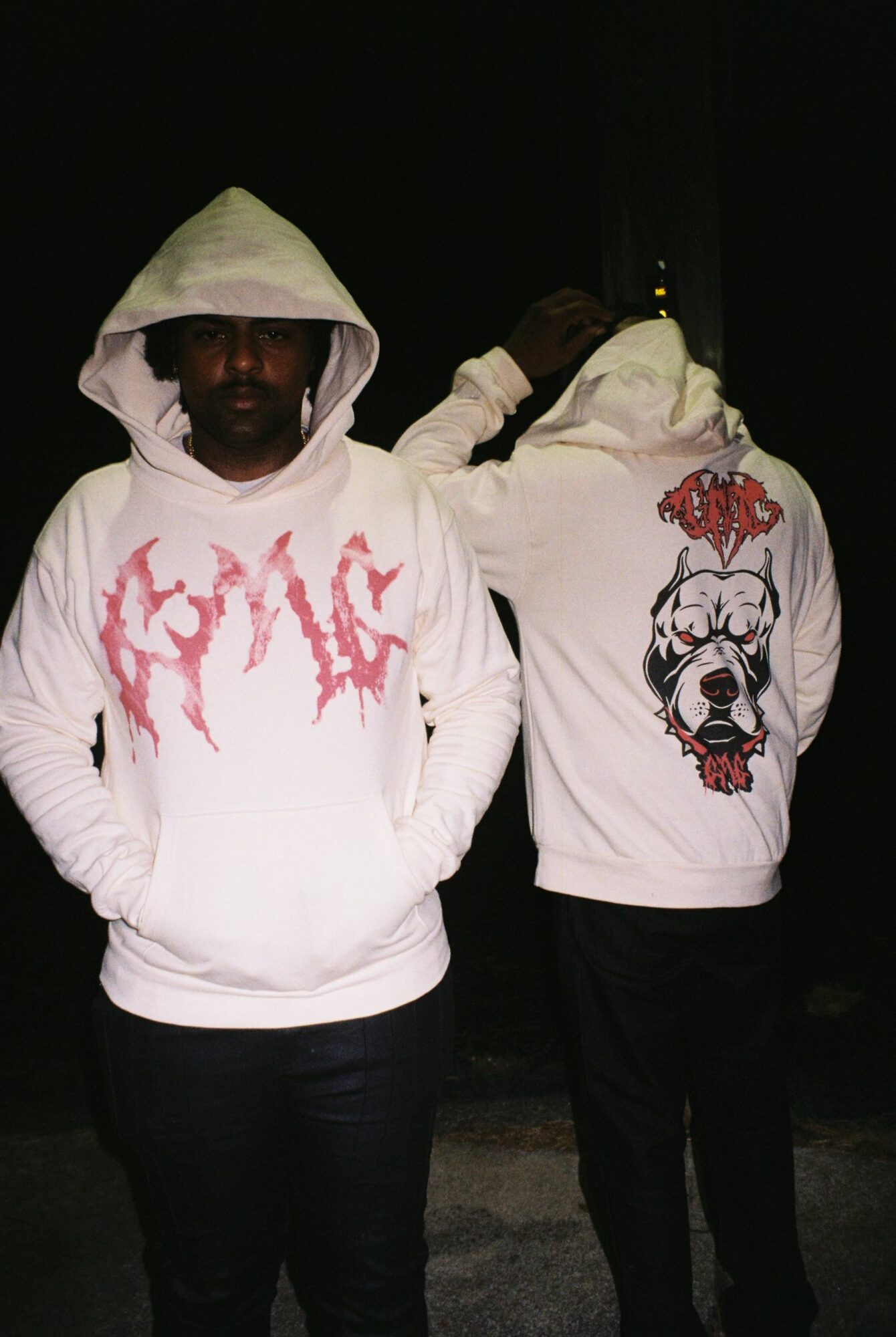 Two people wearing white hoodies with graphic designs, standing in a dark environment, one with a hood over their head.