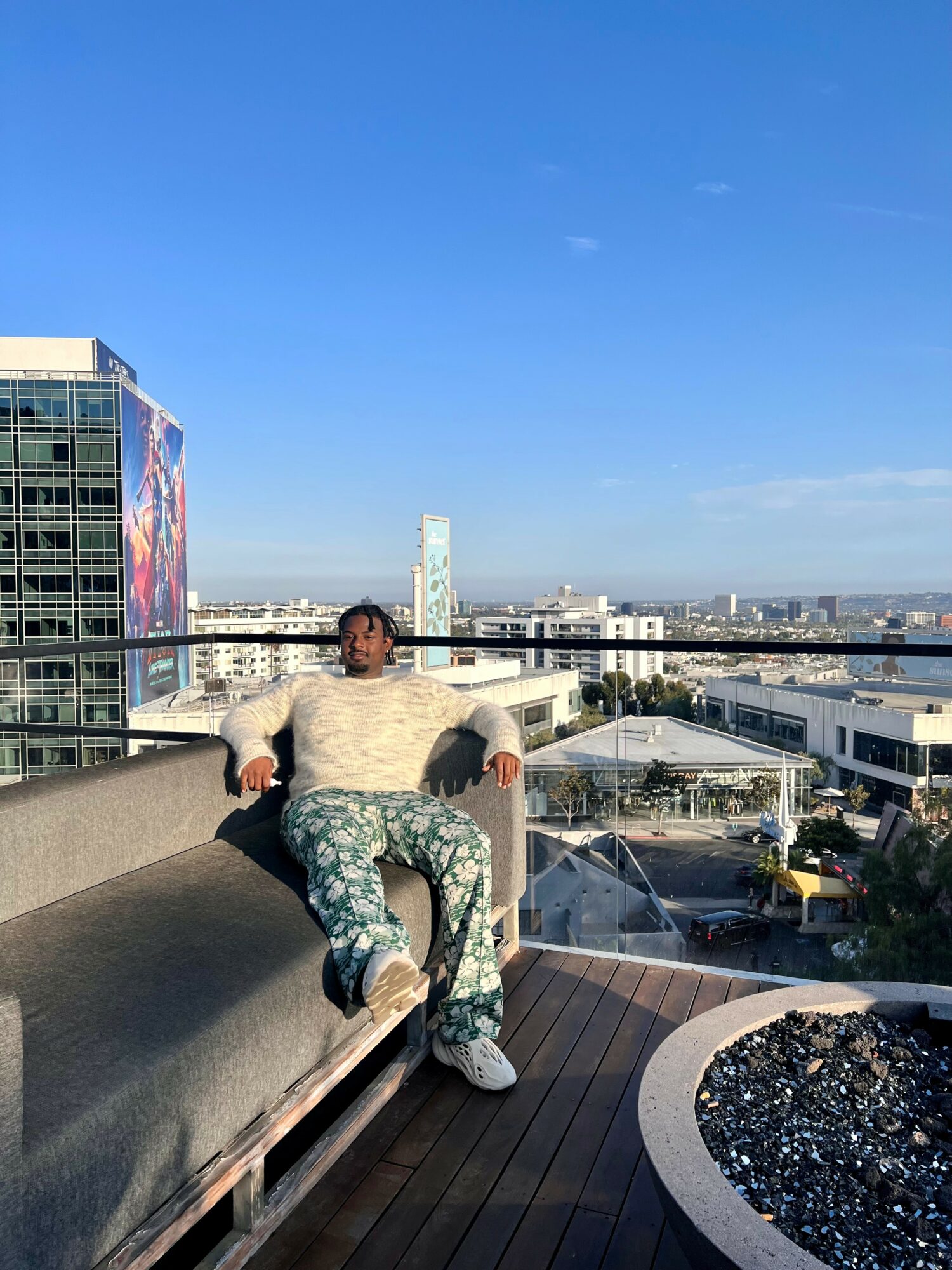 Person sitting on a rooftop ledge with cityscape and blue sky in background.